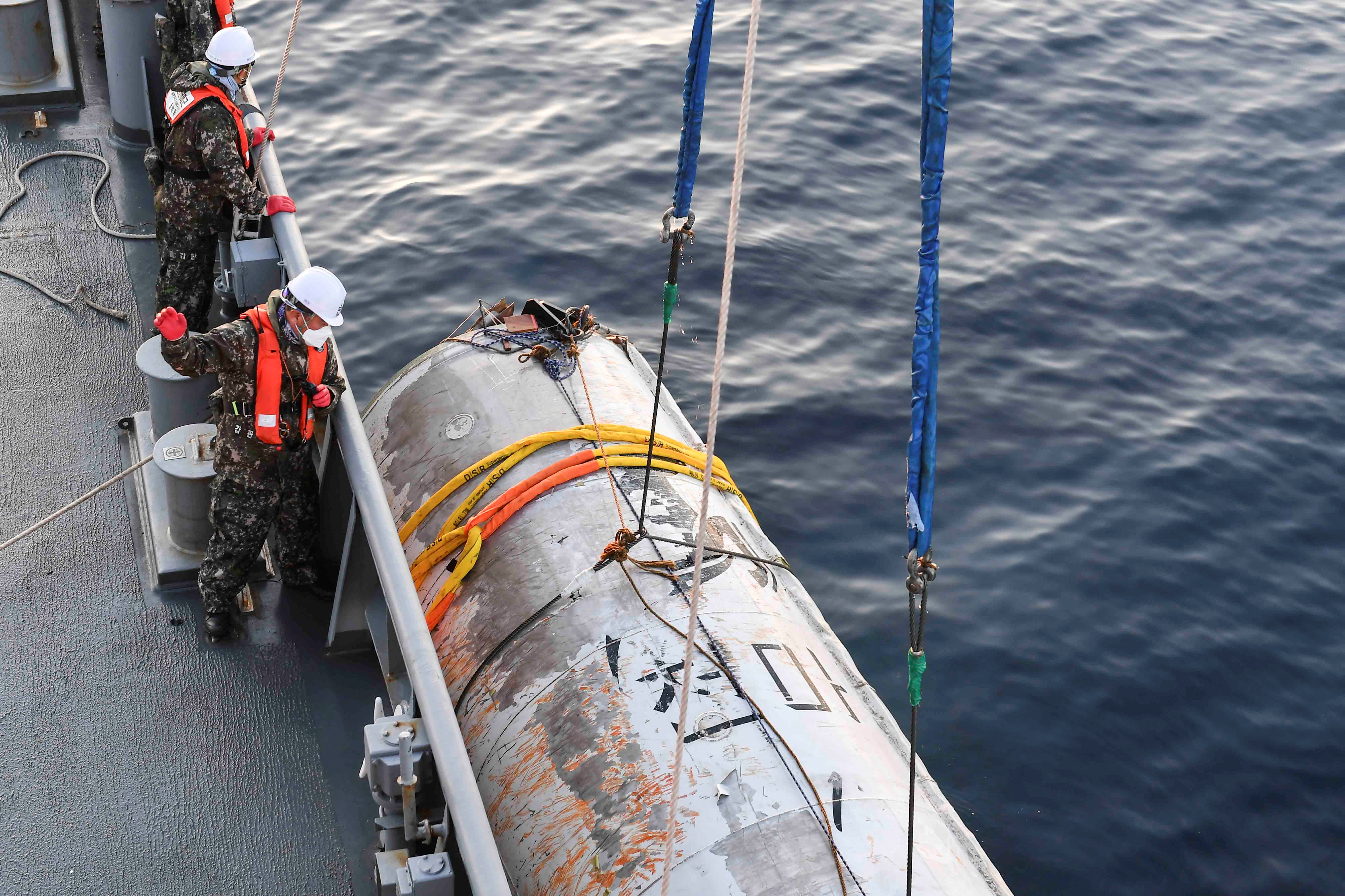 South Korean sailors look down from the deck at what is thought to be a part of a space launch vehicle from North Korea. The wreckage is a large white and silver cylinder with writing in Korean down the middle