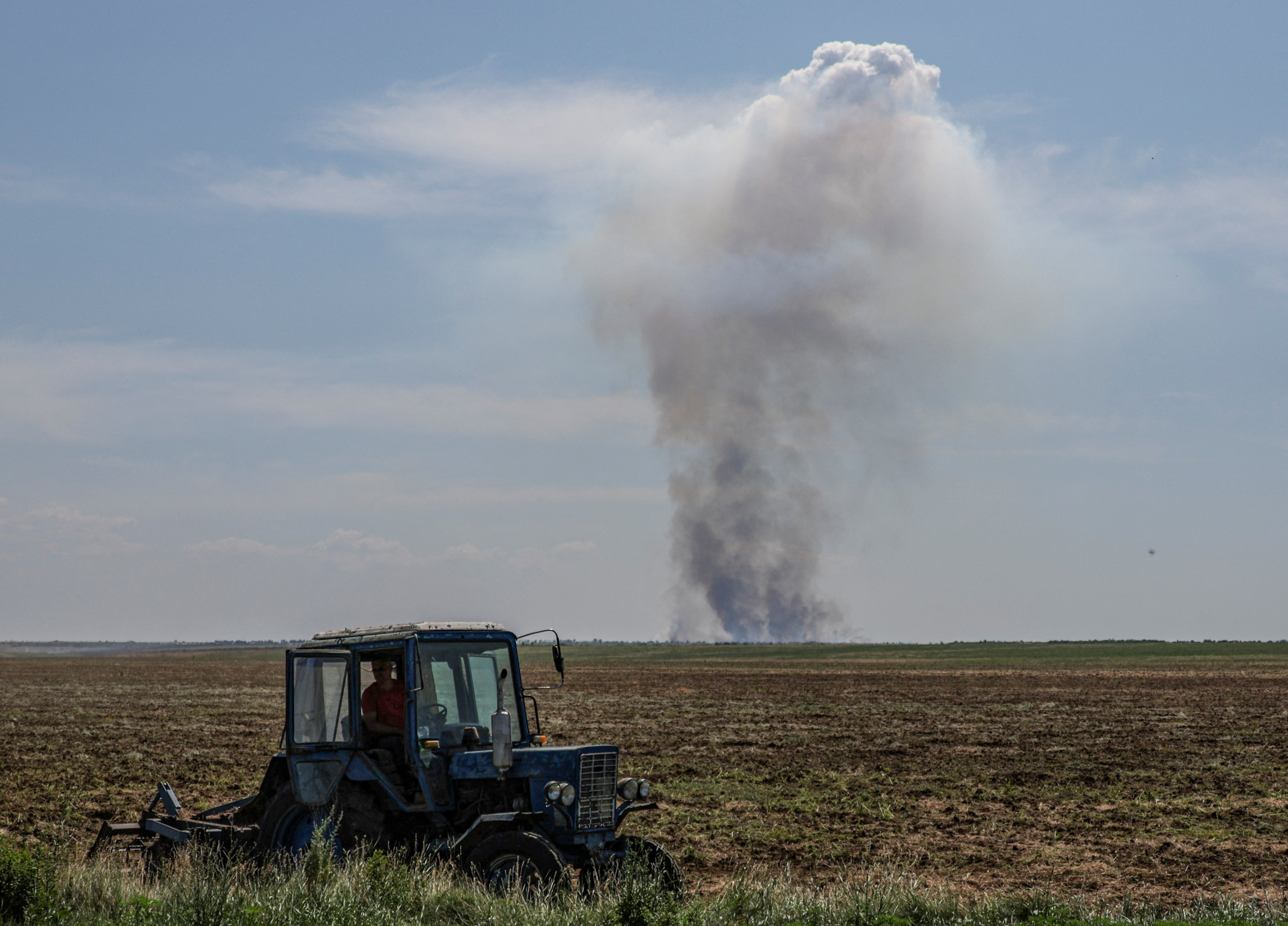A farmer driving a tractor in a vast field. A column of smoke is rising on the horizon after a military attack