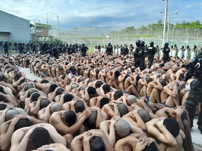 Prisoners, with shaven heads, sit stripped to the waist on the floor, as armed guards look on.