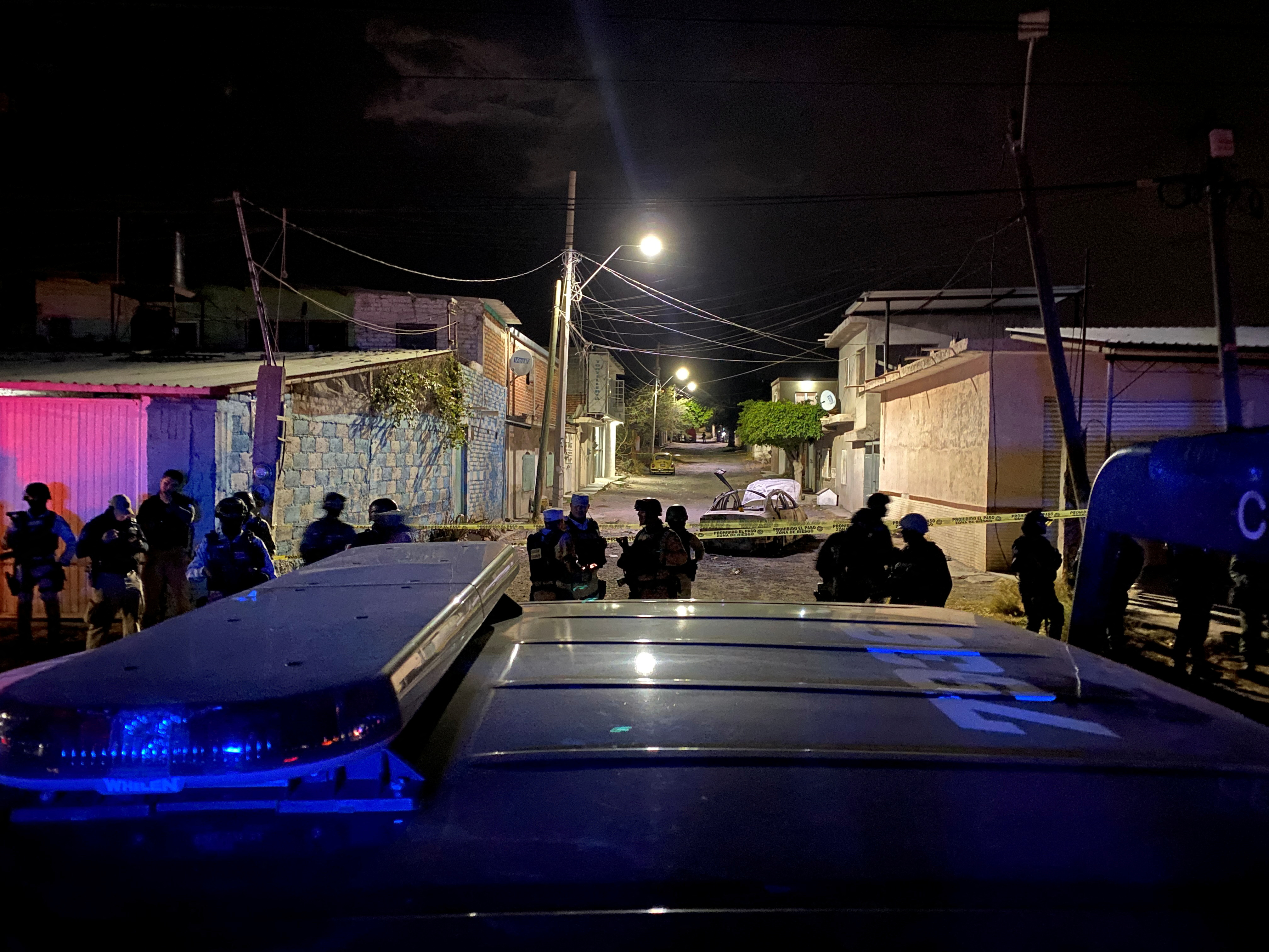 From over the top of a police van, a crime scene is seen, with law enforcement officers surrounding a burned-out car.