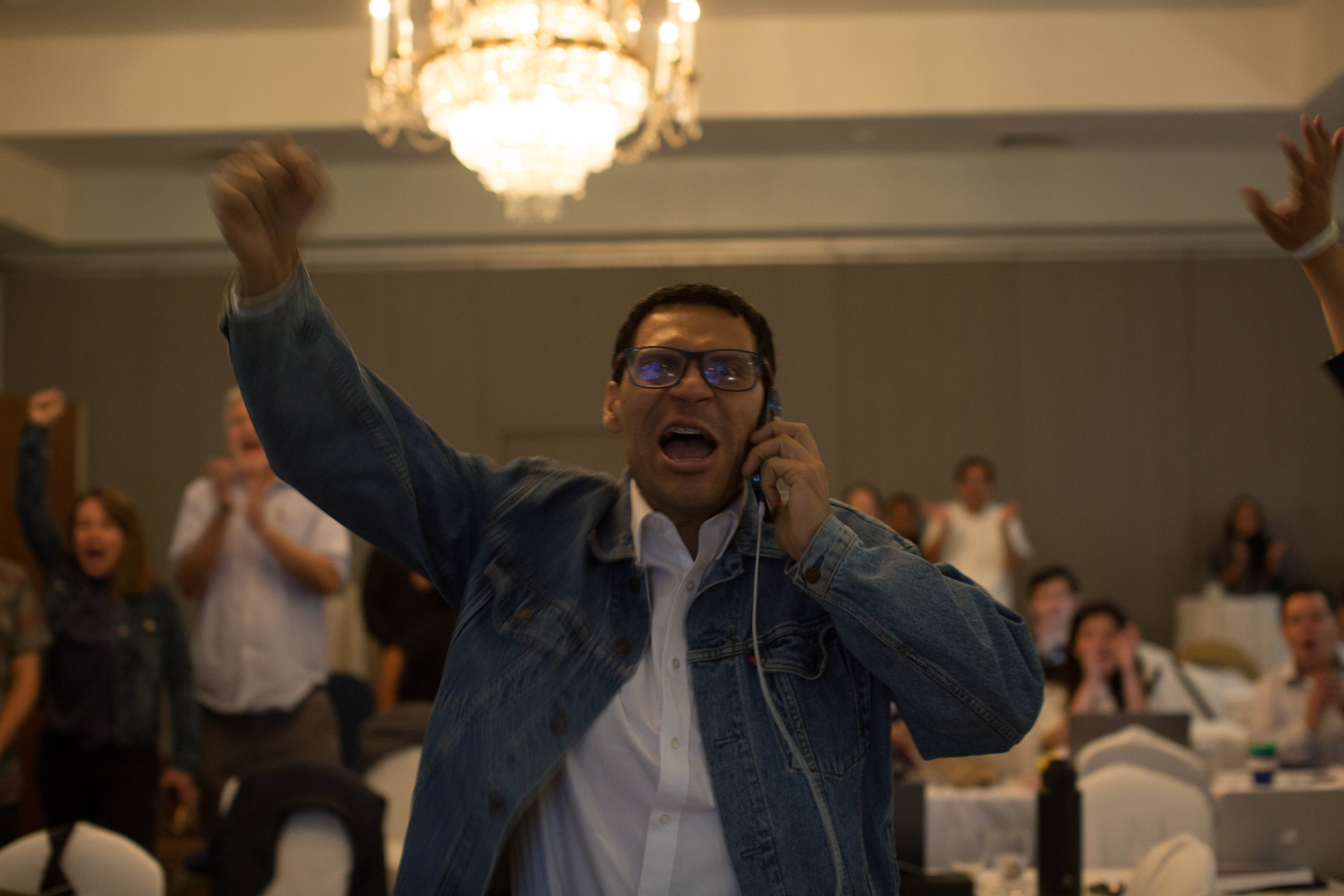 A man in glasses and a jacket raises a fist in the air triumphantly, as he holds a phone to his ear.