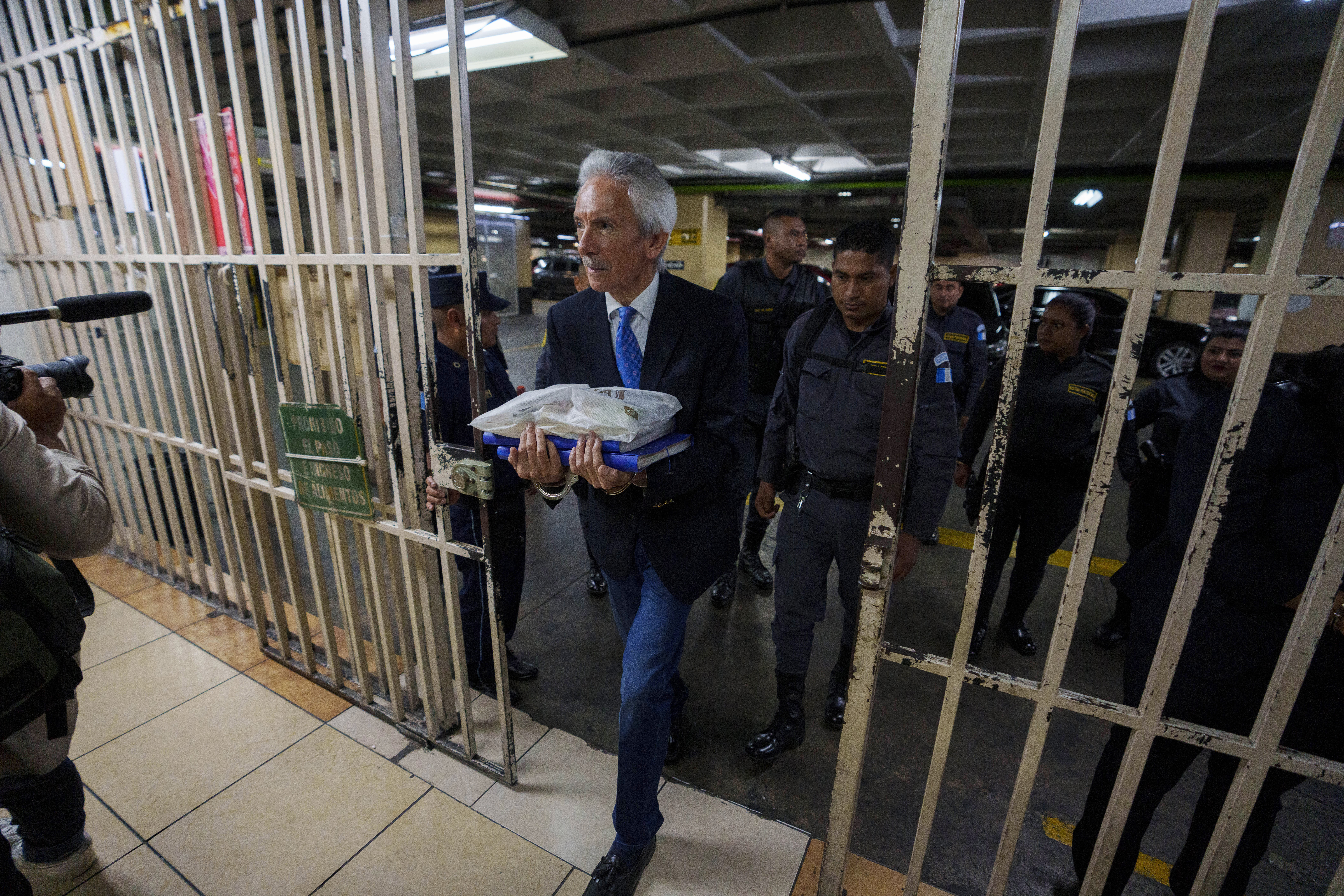 A wall of bars is opened, to allow Jose Ruben Zamora to pass, his hands in cuffs and files in his arms. Police can be seen behind him.