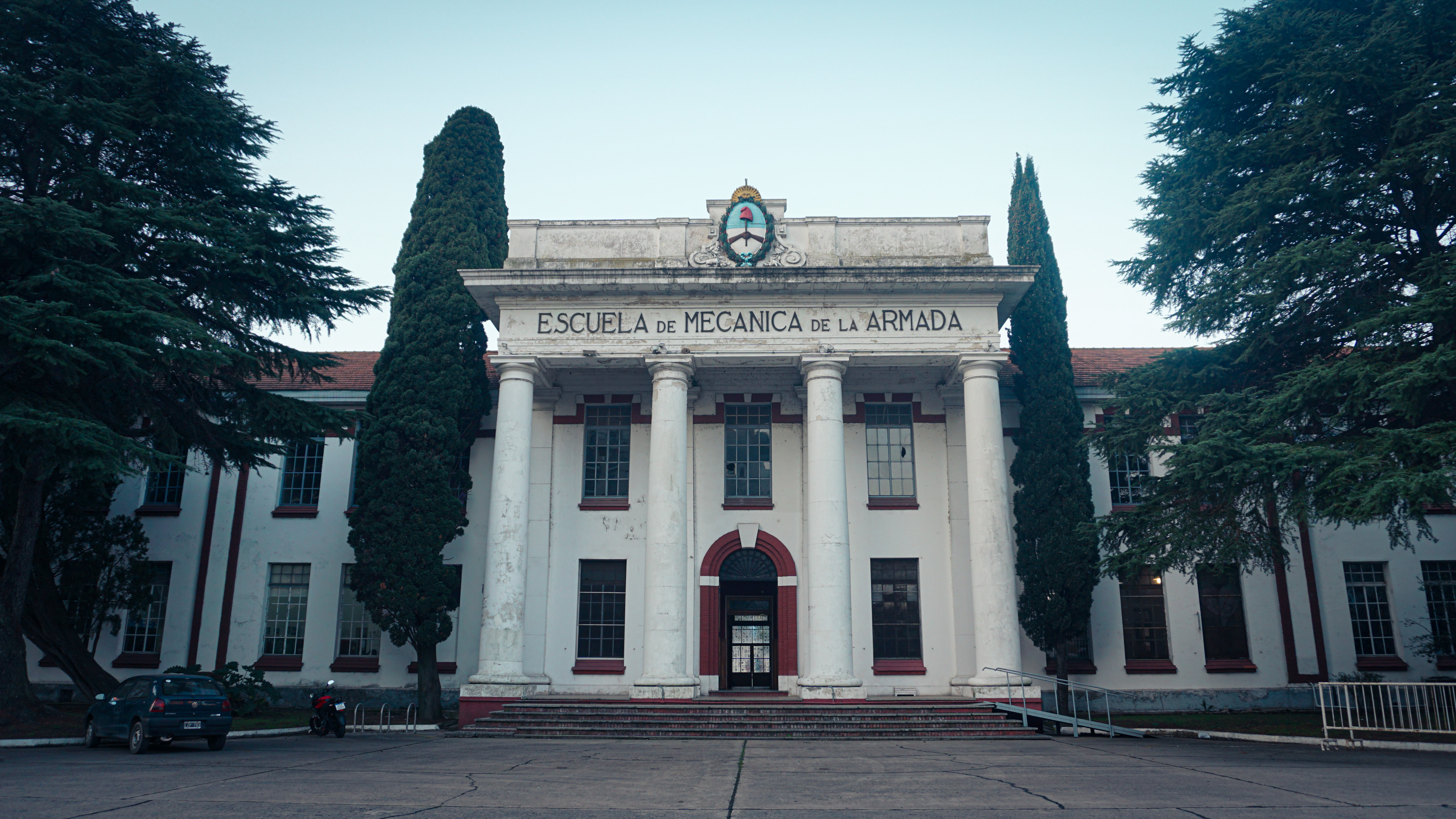 A white two-story building with four columns, in Greco-Roman style, is seen from the sidewalk.
