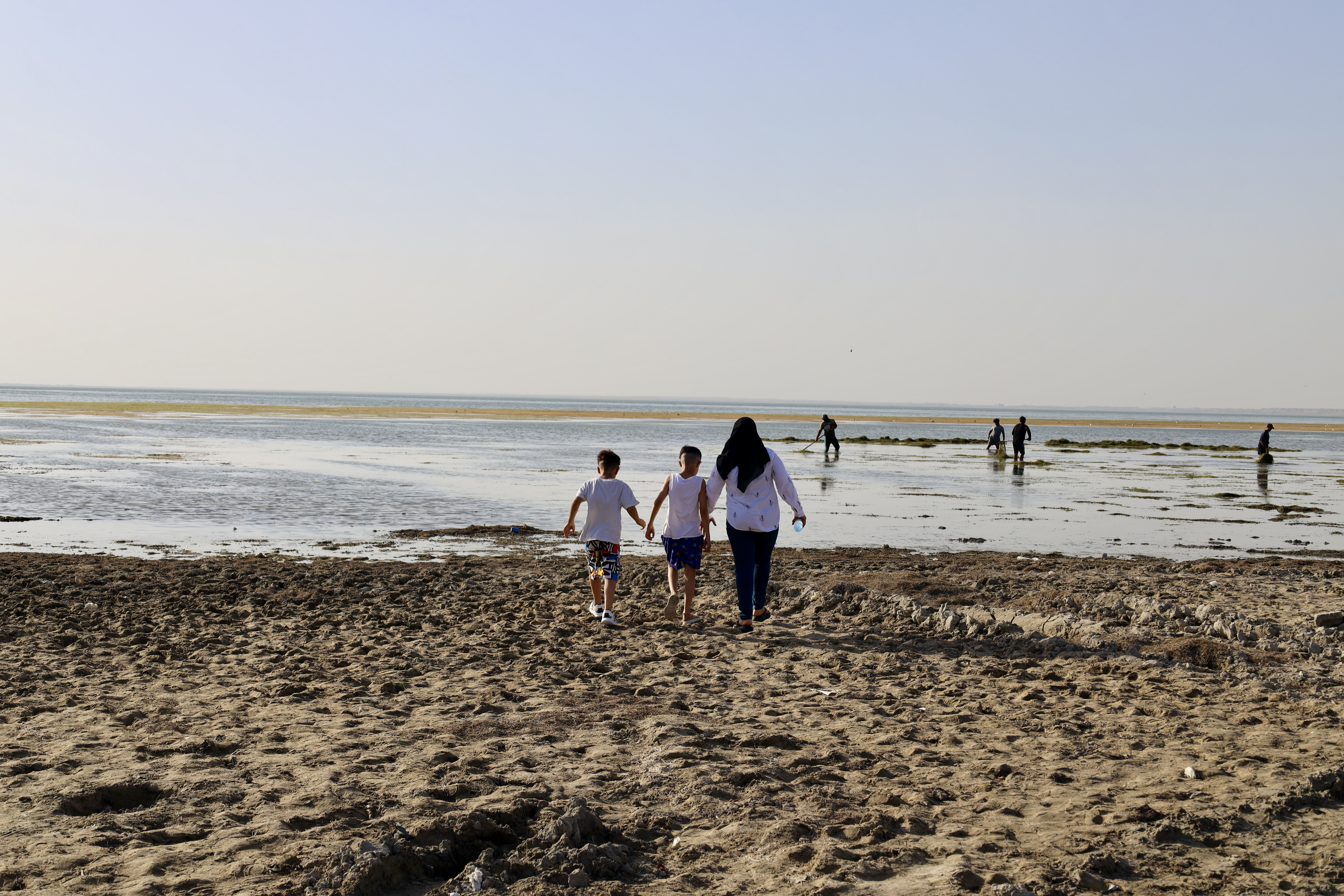 Noor Haddi, 35, and her two young sons venturing towards the distant water of the Habbaniyah Lake resort. In front of them, day labourers earn a daily wage by cleaning natural debris and trash from the water.