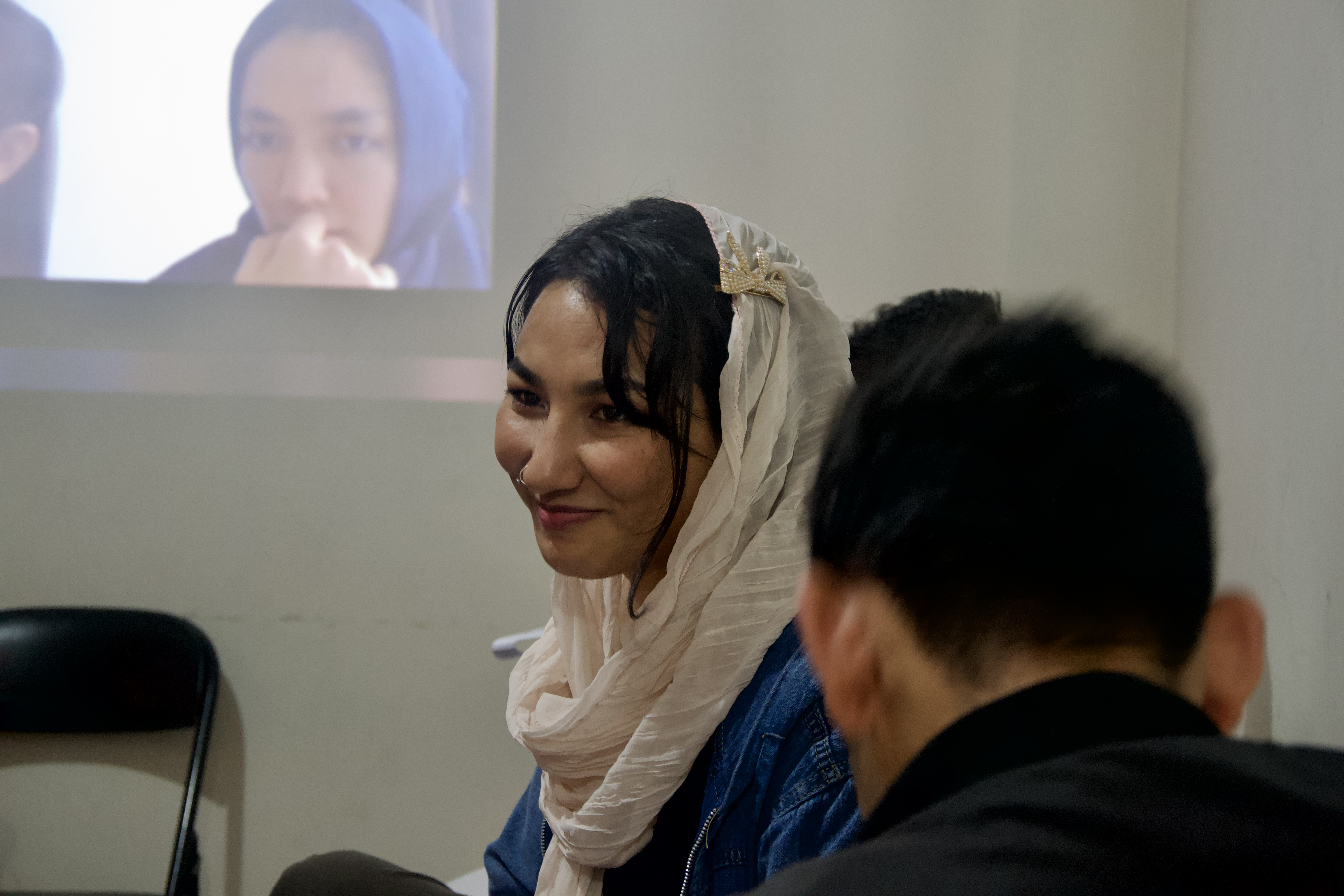 Khatera in her GED class. She has a cream coloured scarf draped loosely around her head. She's smiling.