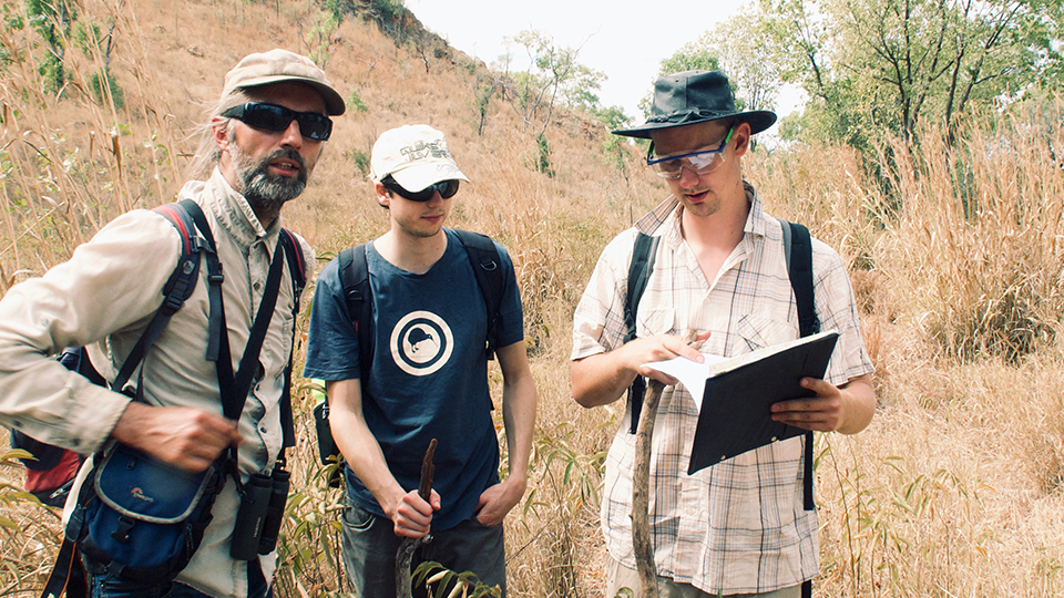 Jochen Brocks and Benjamin Nettersheim, exploring outcrops in northern Australia. They're dressed for hot weather and looking at a file. There is undergrowth and an escarpment behind them