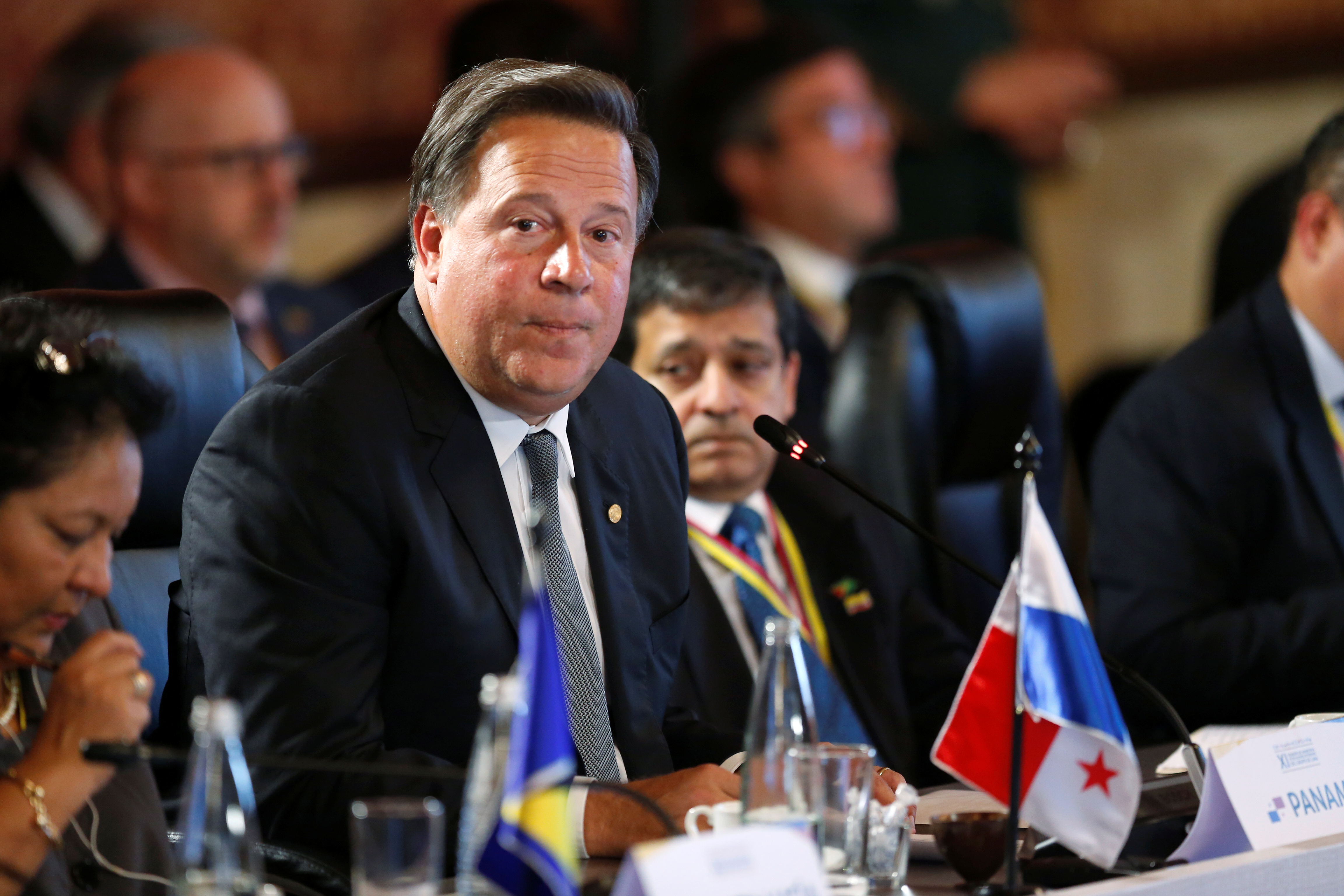 A man leans over a panel table, where a miniature Panamanian flag sits, to speak.