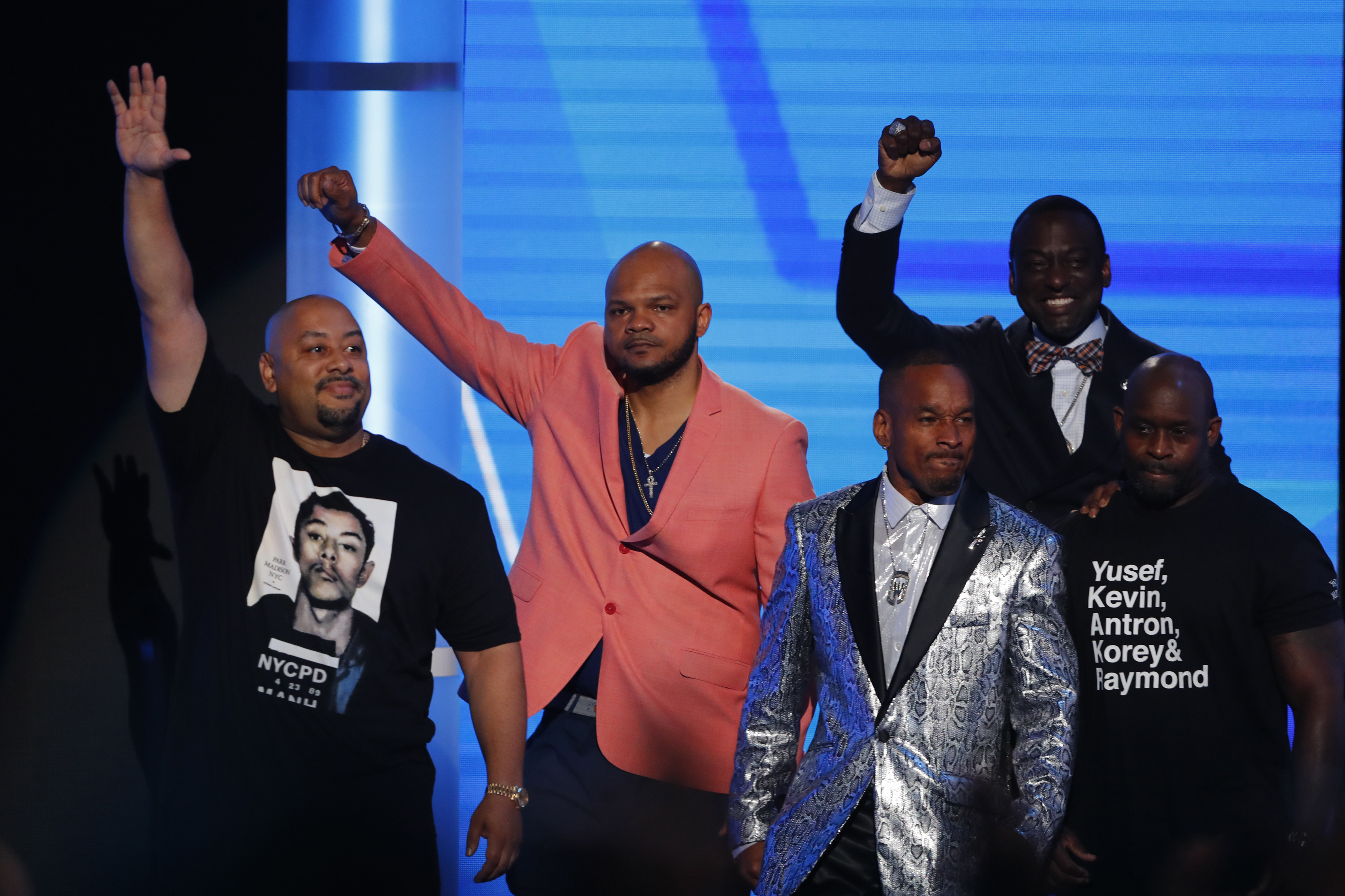 Five men stand on stage, in front of a screen, some of them lifting a fist in solidarity.