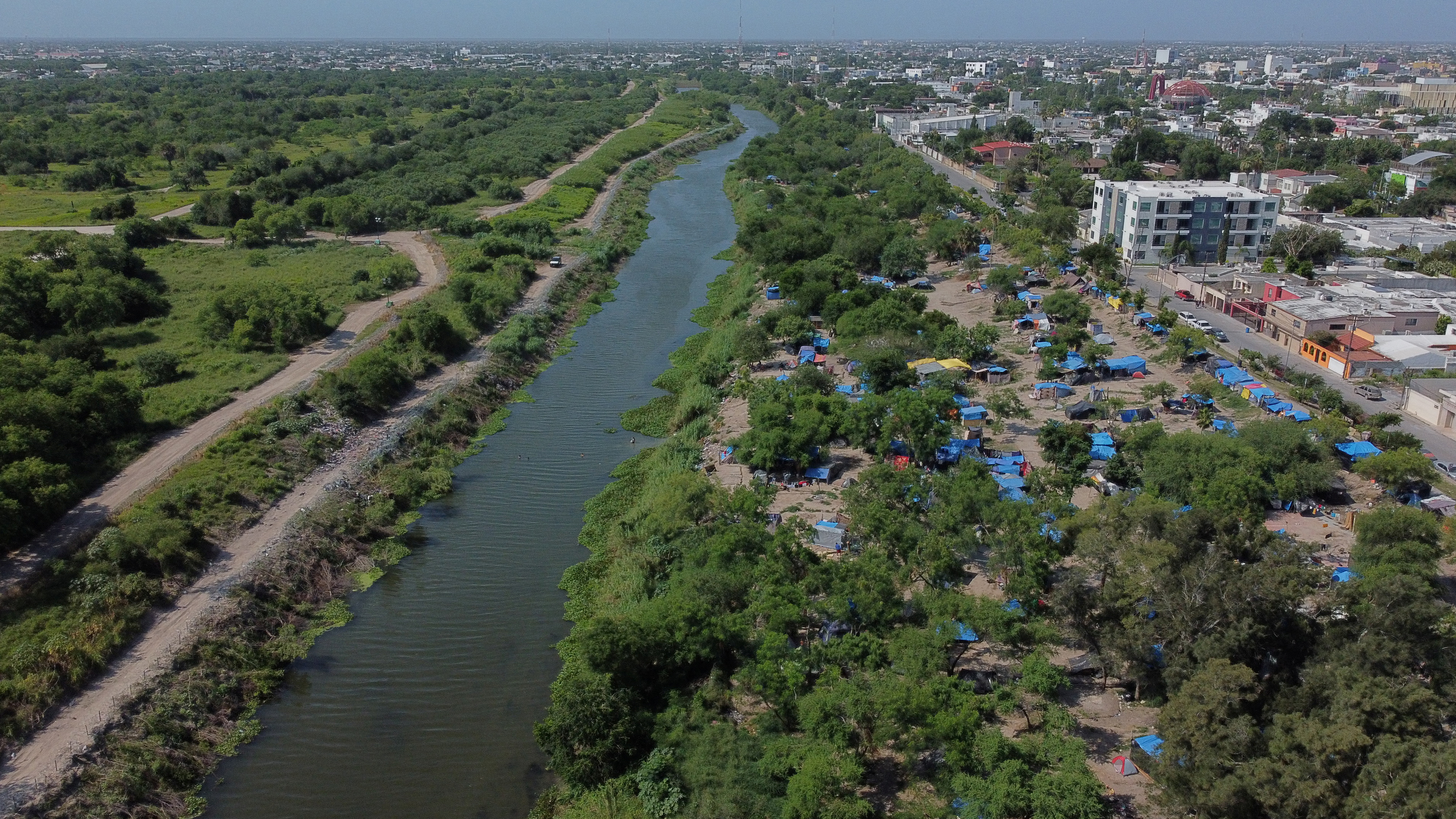 An aerial view of the Rio Grande river, a wide river surrounded by greenery, with a city and makeshift encampment with blue tents visible nearby.
