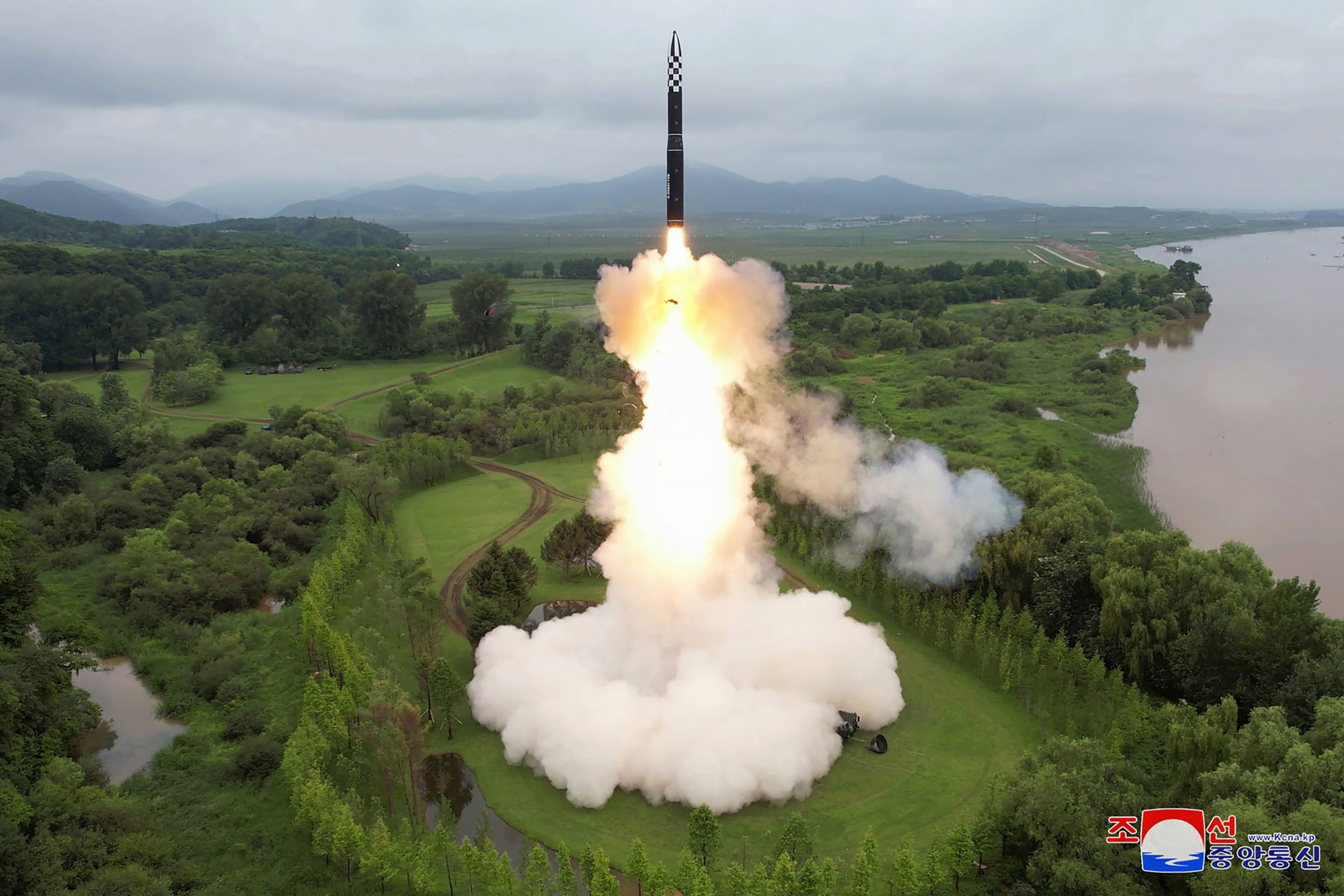 The missile launching into the air. There are lots of smoke clouds. The area around is green and lush and there is water to the right