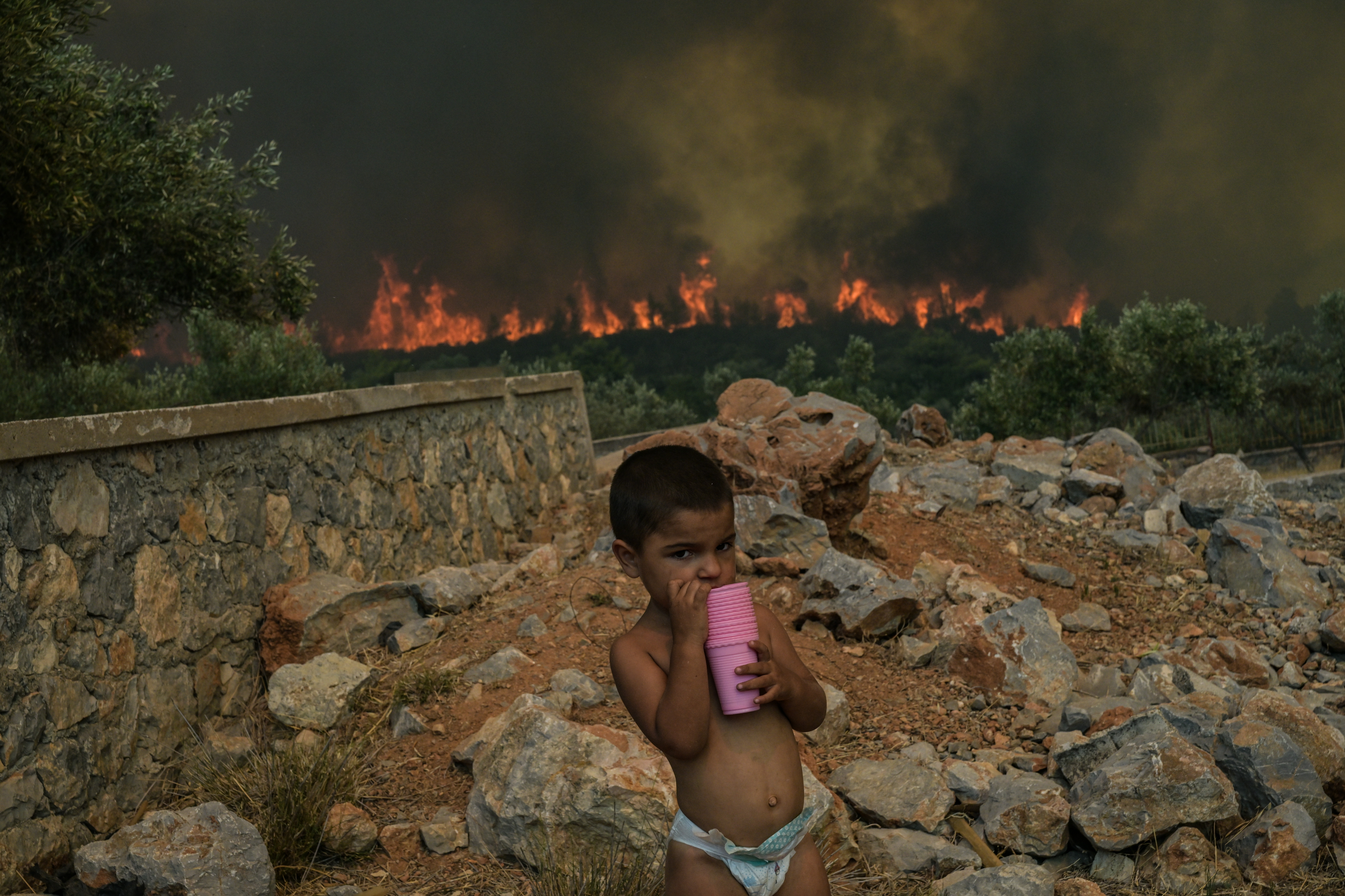 A child stands at the yard of his house as a wildfire burns, in the village of Agios Charalampos, near Athens, on July 18, 2023. - Europe braced for new high temperatures on July 18, 2023, under a relentless heatwave and wildfires that have scorched swathes of the Northern Hemisphere, forcing the evacuation of 1,200 children close to a Greek seaside resort. Health authorities have sounded alarms from North America to Europe and Asia, urging people to stay hydrated and shelter from the burning sun, in a stark reminder of the effects of global warming. (Photo by Aris MESSINIS / AFP)