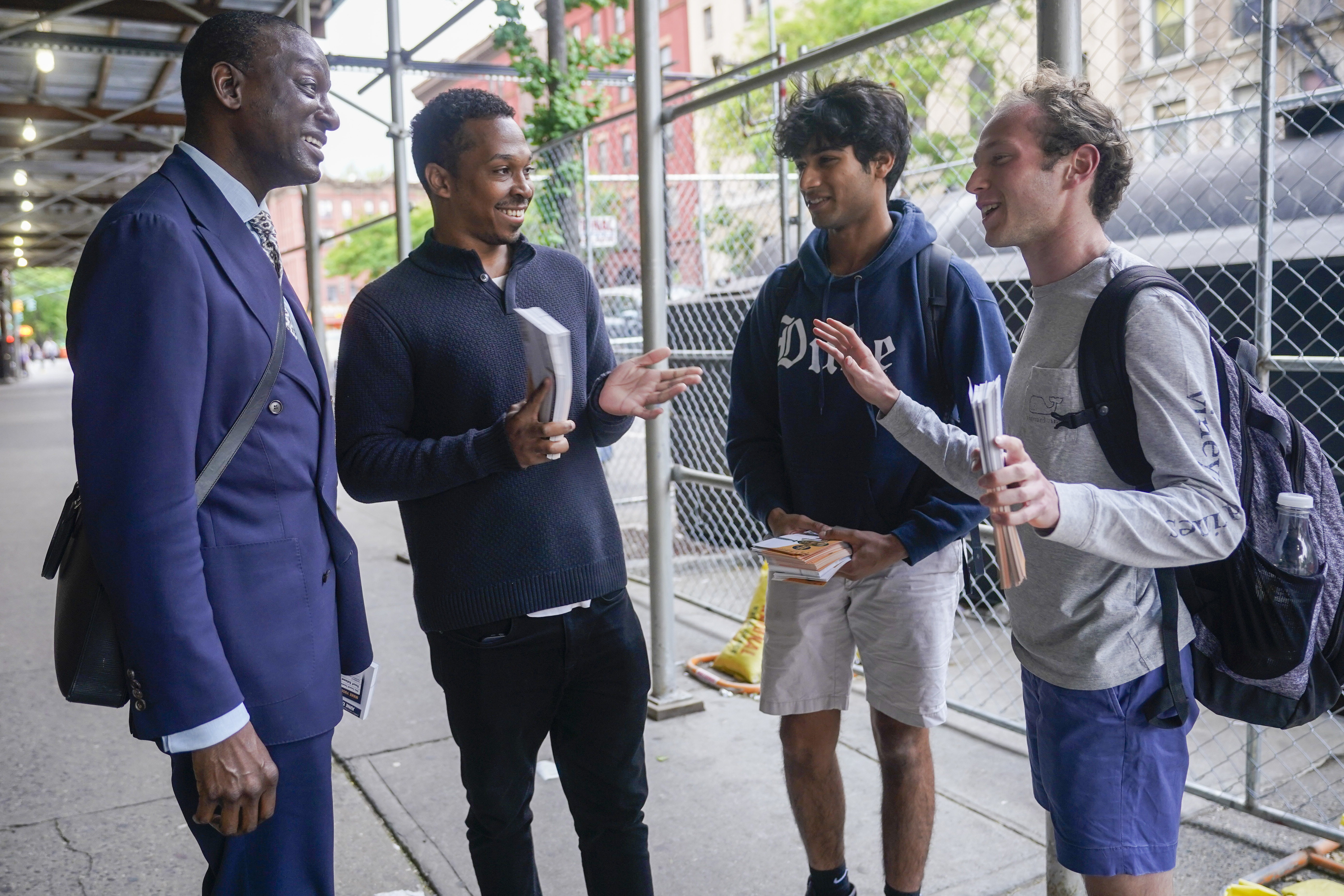 A tall man in a navy suit speaks to young volunteers — in hoodies, pullover sweaters, shorts and backpacks — under some scaffolding outside in New York City.