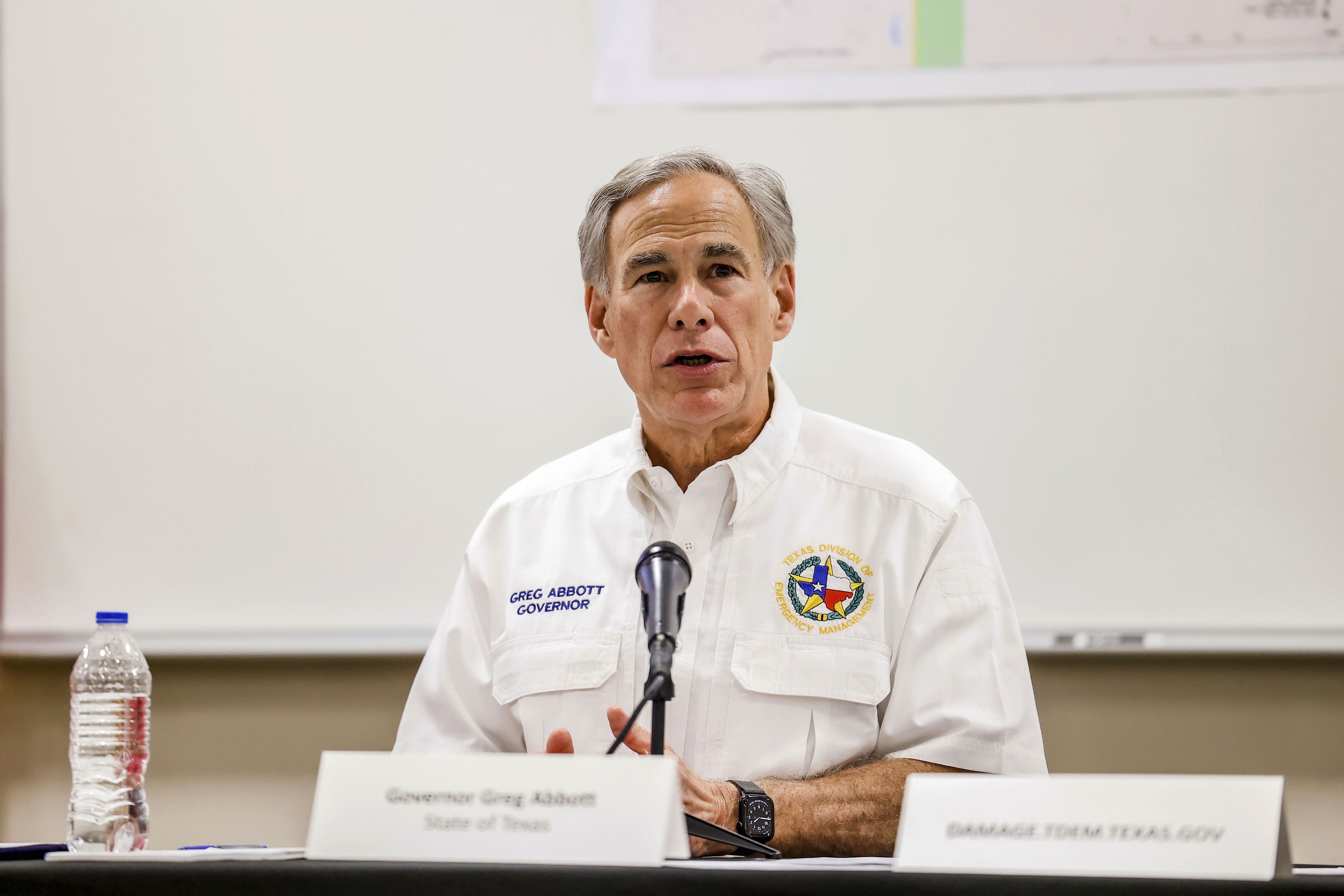 Governor Greg Abbott, dressed in a white uniform, sits at a panel table with a microphone in front of him.