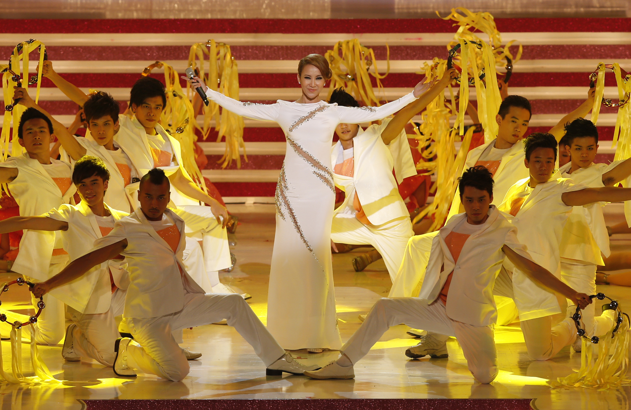 Coco Lee performing on stage in Hong kong. She is wearing a white dress and is surrounded by dancers in white suits holding tambourines with yellow ribbons, She is taking a bow.
