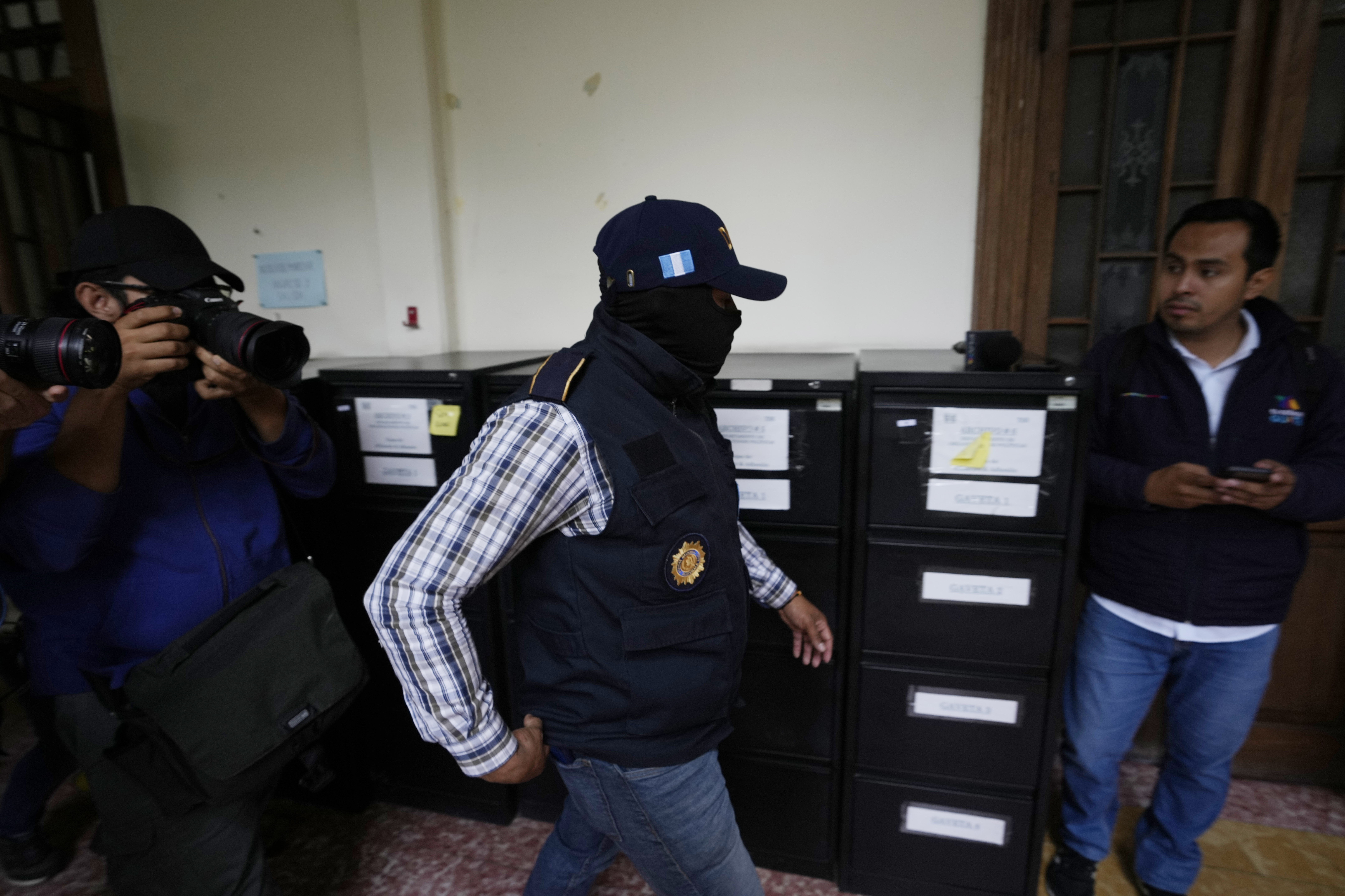 A man in a dark vest and baseball cap enters an office with filing cabinets.
