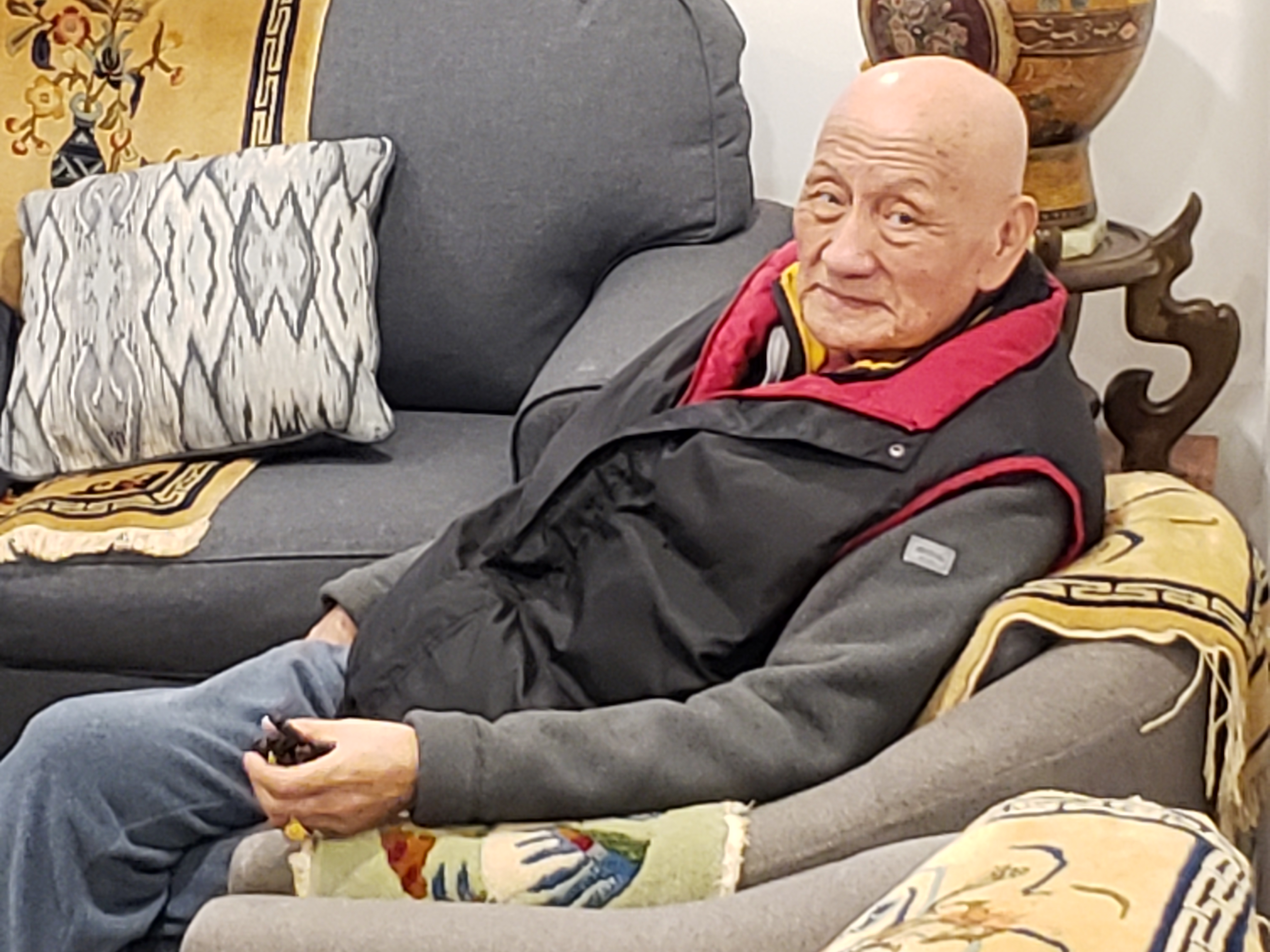 Tsultrim pictured in New York, He is seated ona grey sofa and wearing a fleece-style jacket with a body warmer. He is bald.