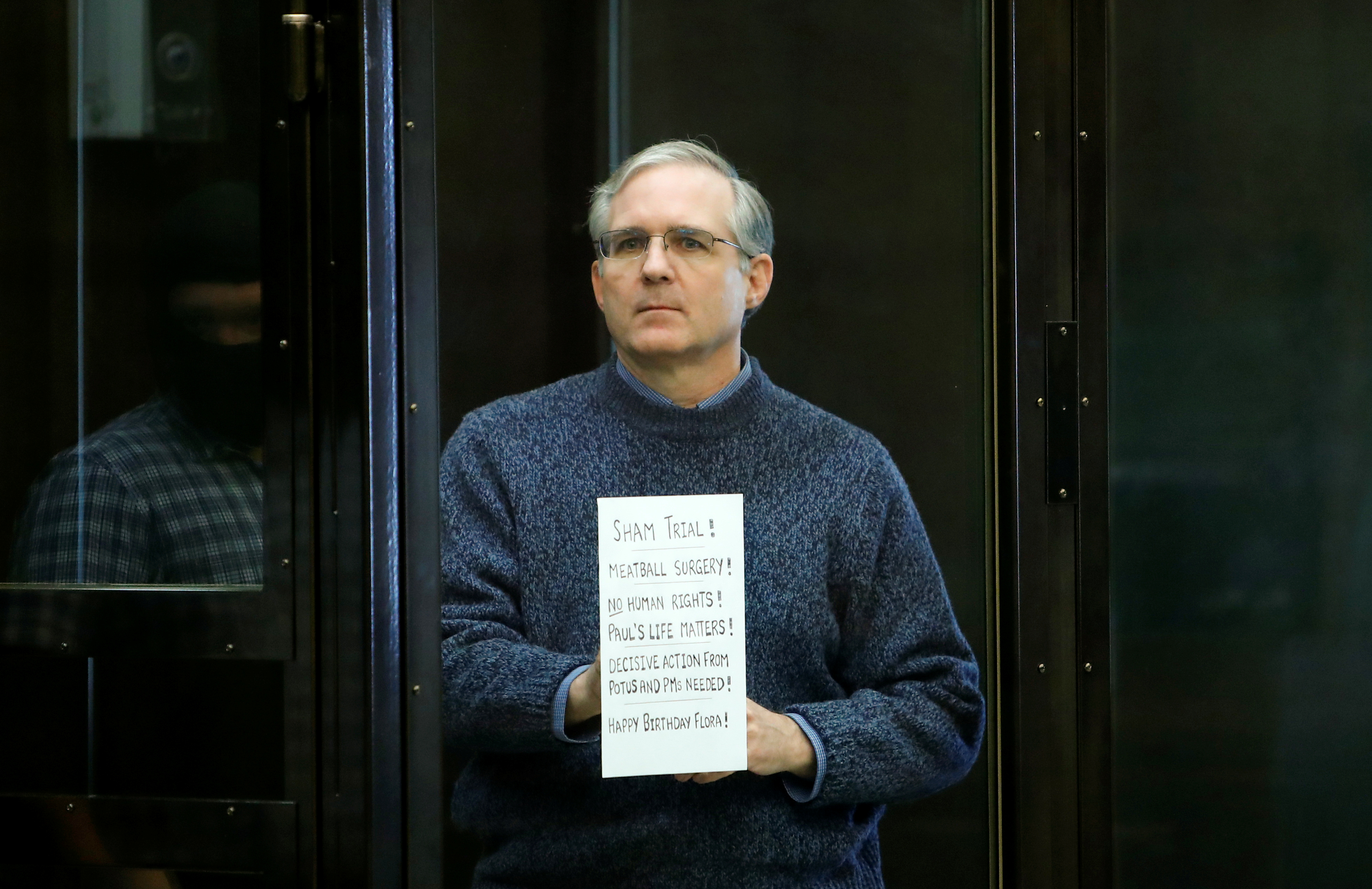 A man, Paul Whelan, stands in a glass box holding a sign that reads, "Sham trial! Meatball surgery! No human rights! Paul's life matters! Decisive action from POTUS and PM needed! Happy birthday, Flora!"