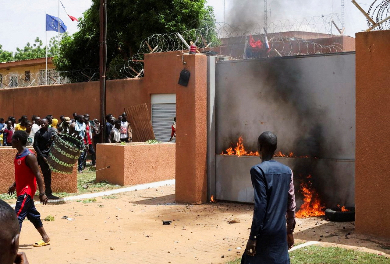 A person passes outside the walls of an embassy compound, as a fire burns at its base.