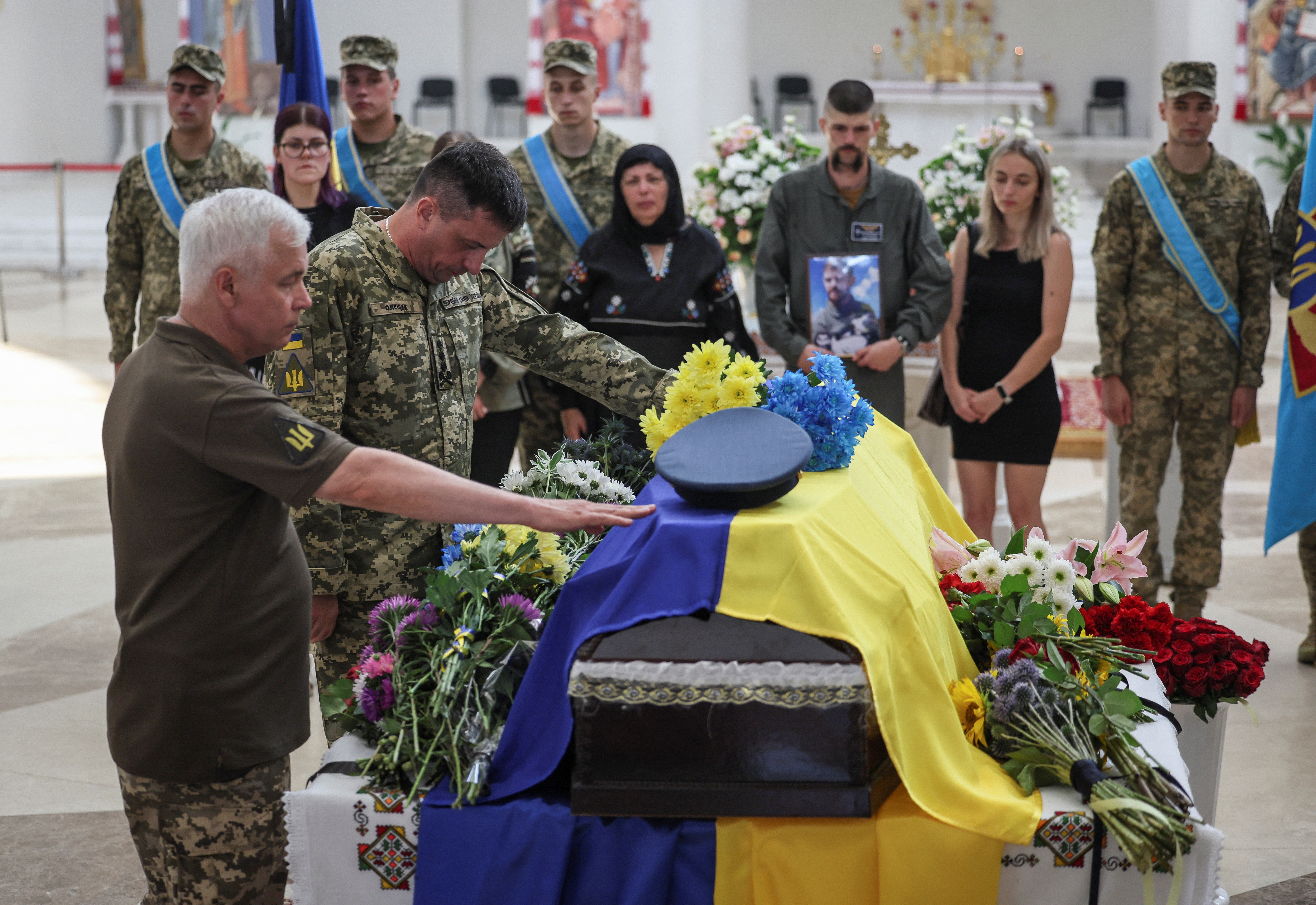 Ukrainian servicemen around the coffin of fighter pilot Andriy Pilshchykov. They have their heads bowed. The coffin is covered in a Ukrainian flag. His air force cap is on top among flowers.