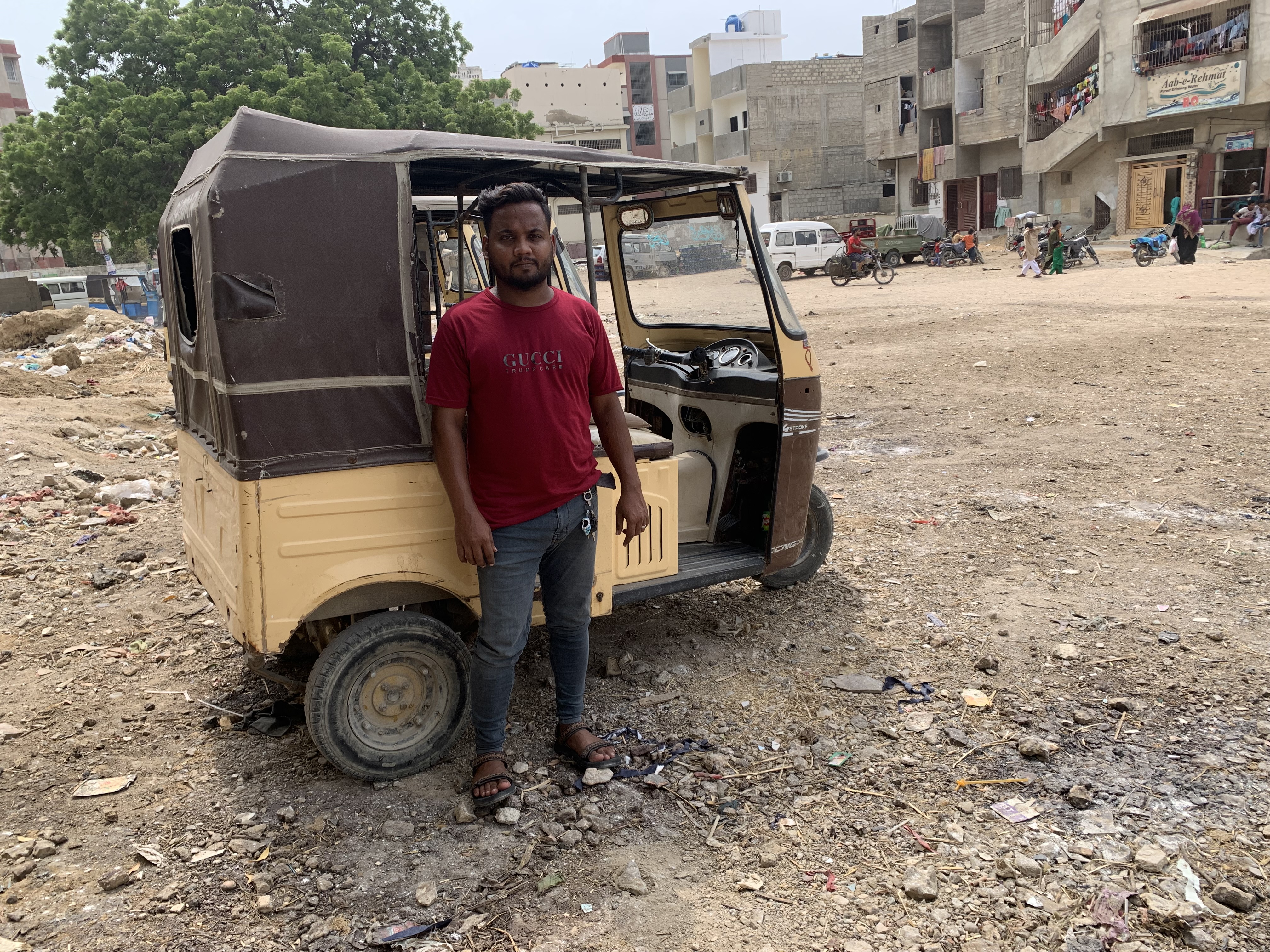 A photo of Faisal standing next to his rickshaw with shops in the background.