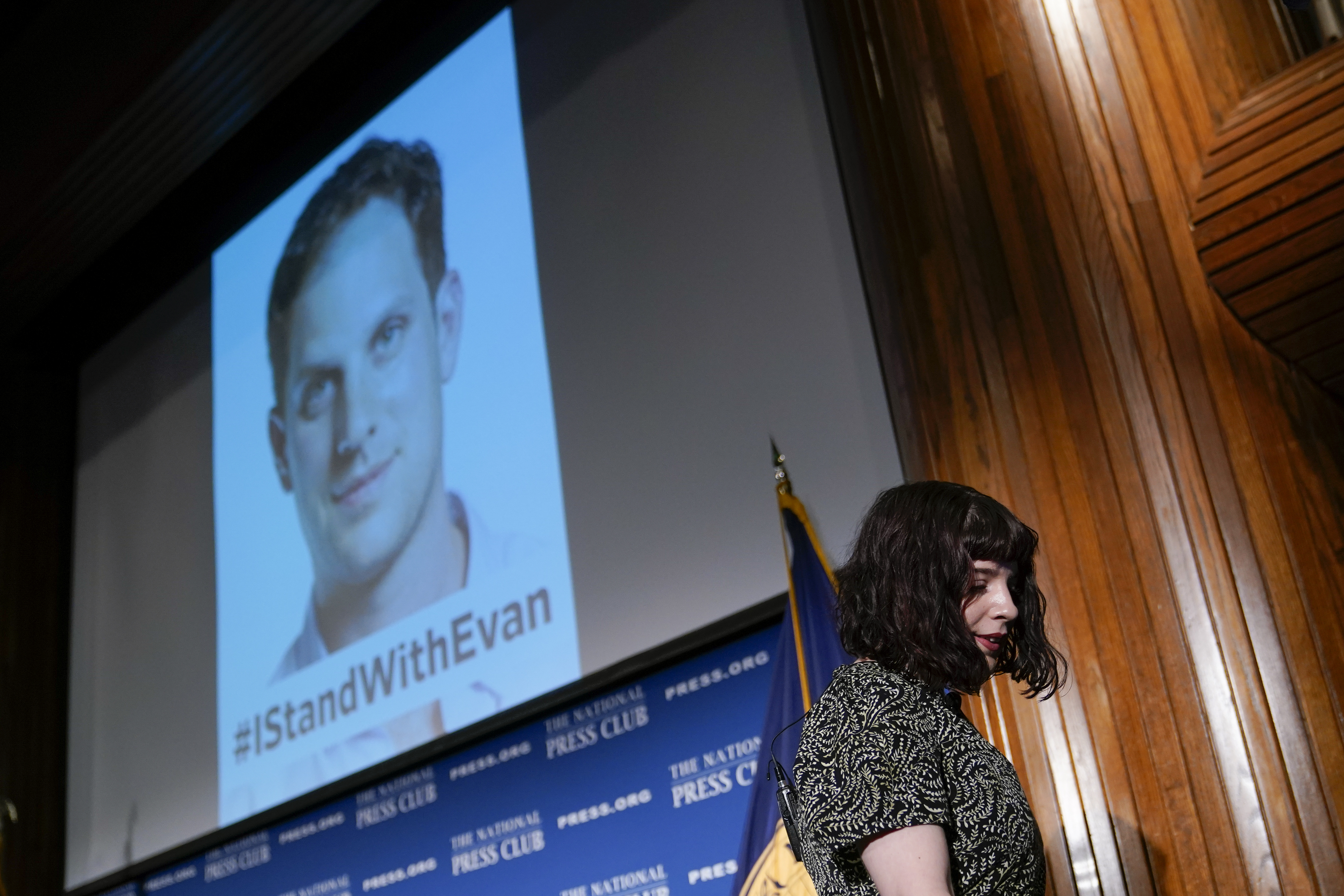 A woman, Danielle Gershkovich, steps down from a stage, above which a screen projects an image of her brother.