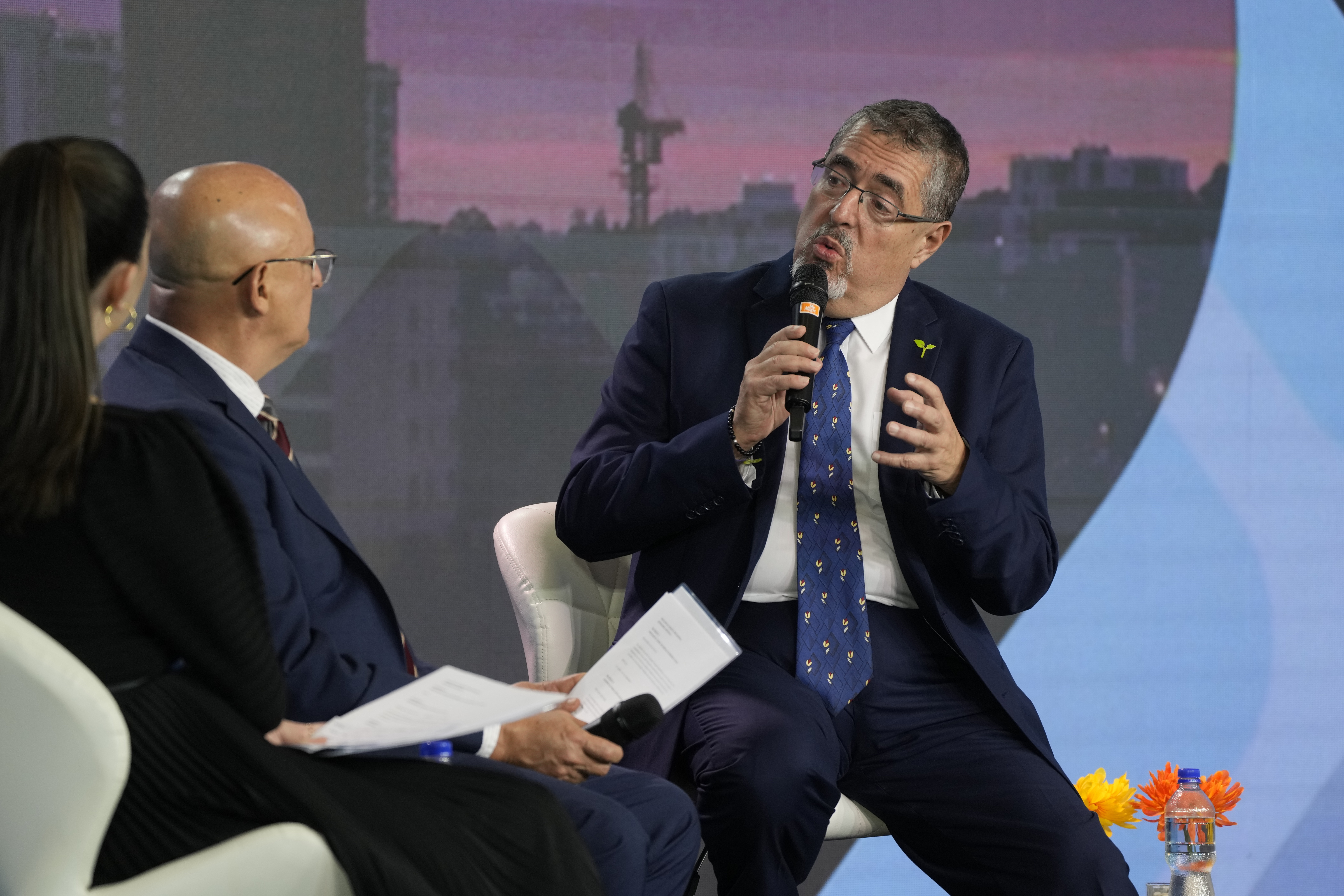 A man in a dark suit and glasses, Bernardo Arevalo, sits on a white chair and speaks into a microphone, his arms lifted slightly in gesture. Interviewers sit across from him on the stage.