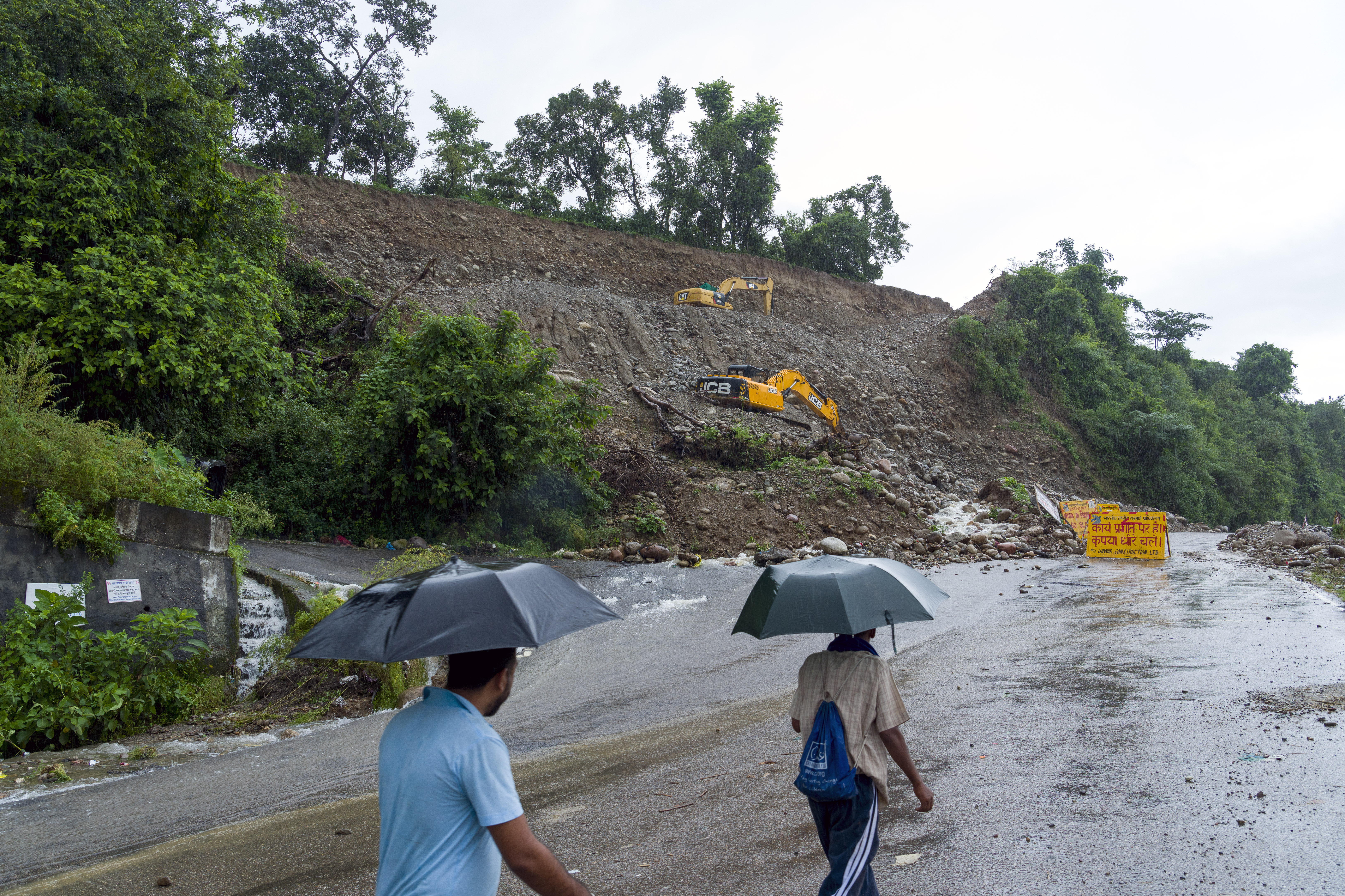 People walk in the rain next to a highway expansion site where heavy rains have disrupted operations and blocked part of the existing road in Dharamshala, India, Monday, Aug. 14, 2023. Heavy monsoon rains triggered floods and landslides in India's Himalayan region