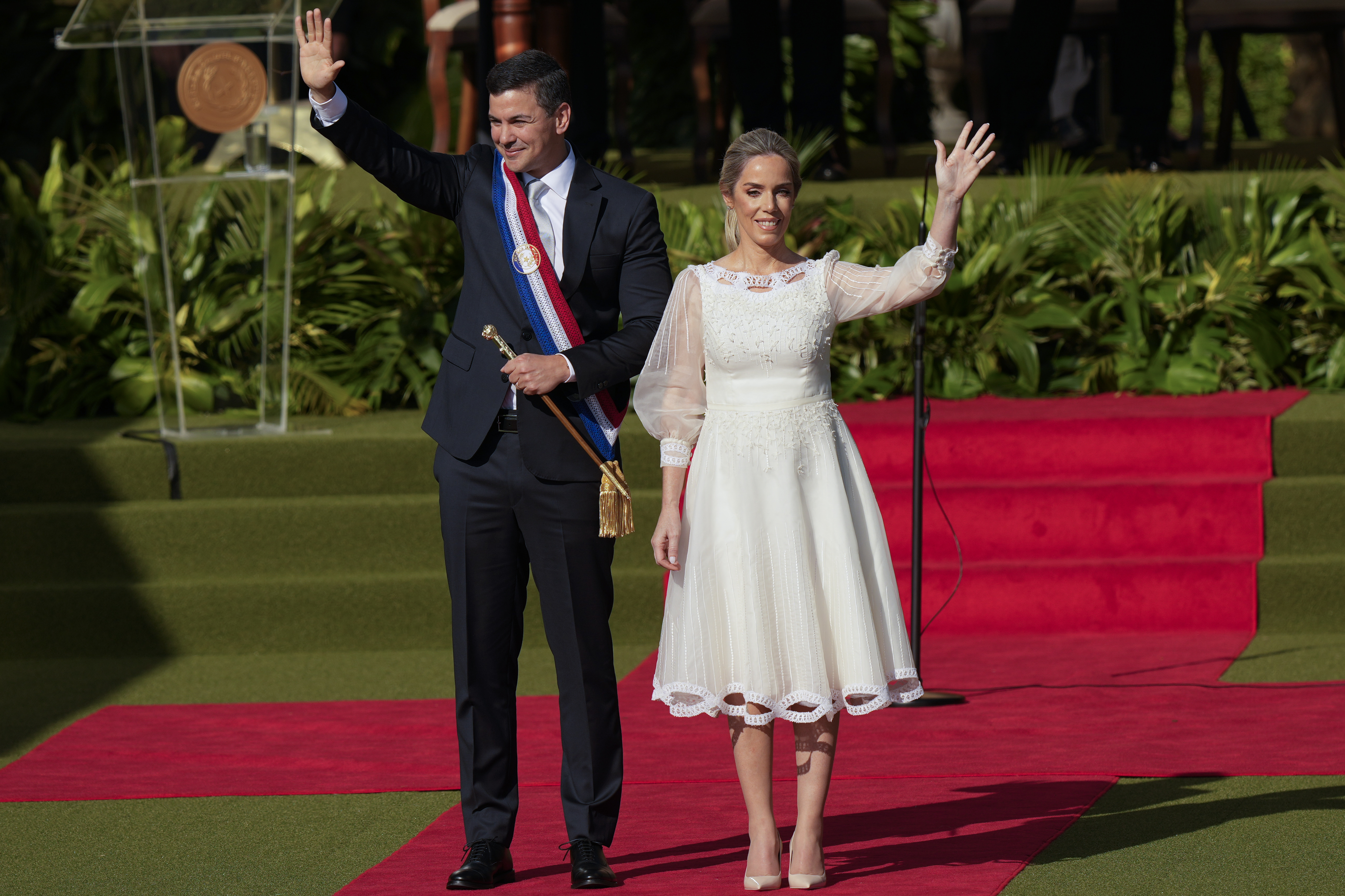 A man in a dark suit and red-white-and-blue sash stands side by side with a woman in a white dress, as they wave from the red carpet.