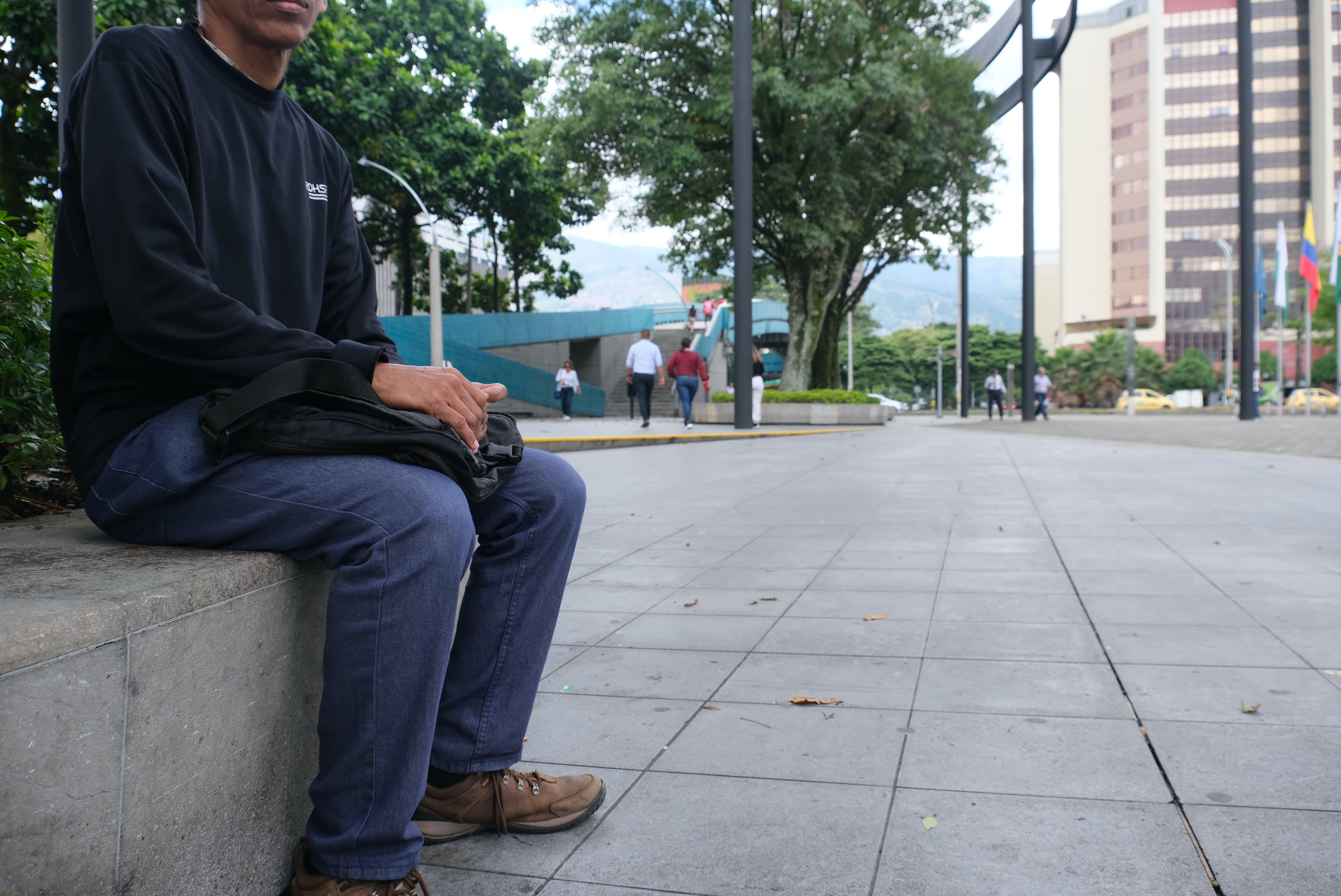 A man — obscured by the edges of the photo — sits on a concrete bench on the sidewalk in Medellin, Colombia. He wears jeans, a long-sleeve pullover and brown sneakers.