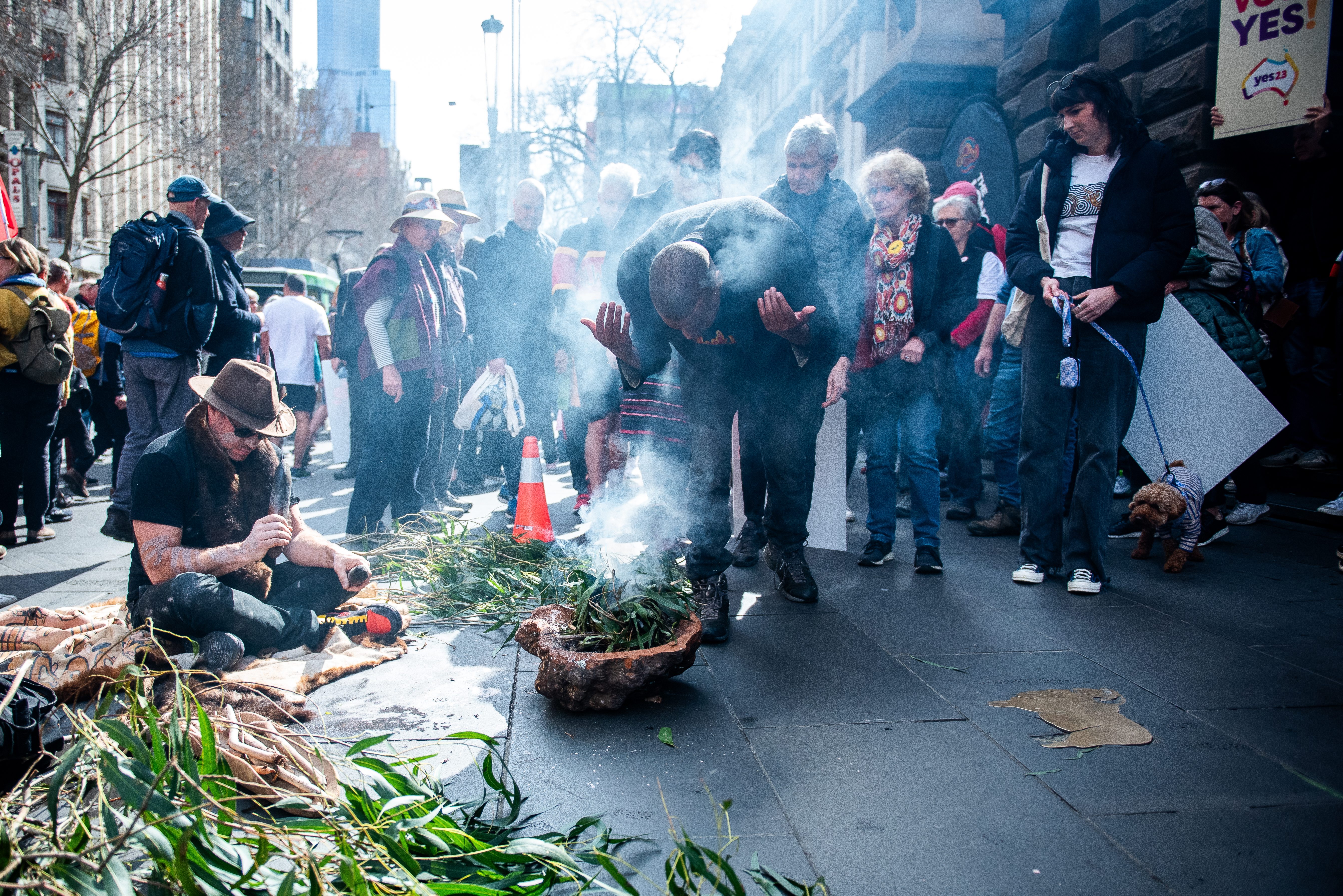 A man conducting a traditional smoking ceremony on the road outside Melbourne Town Hall. He is leaning over a wooden bowl containing green leaves that are giving off smoke. A man to his side is speaking into a microphone. There are people watching the ceremony.