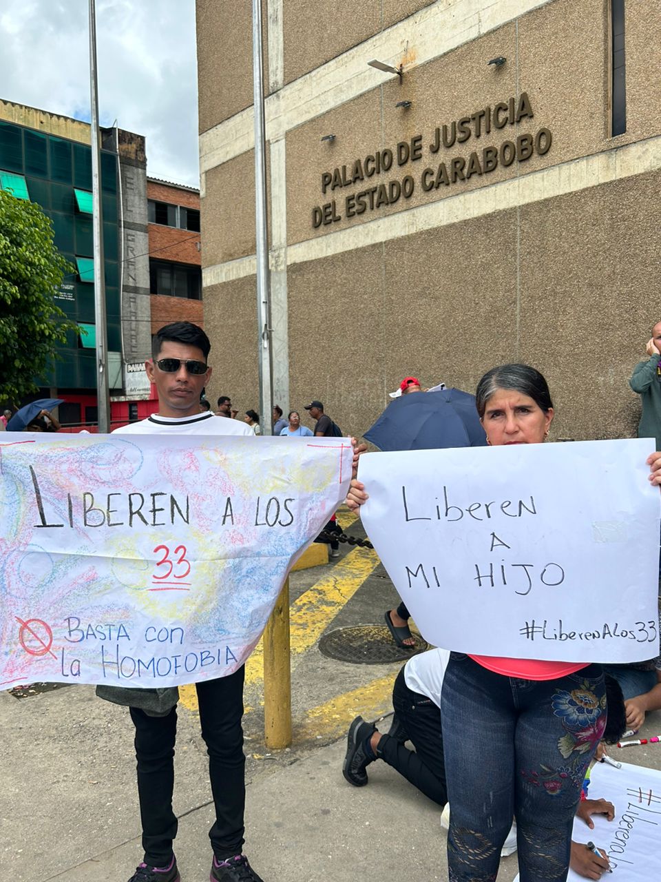 Two people — a young man and a woman — hold up posters with handwritten messages outside of a courthouse.