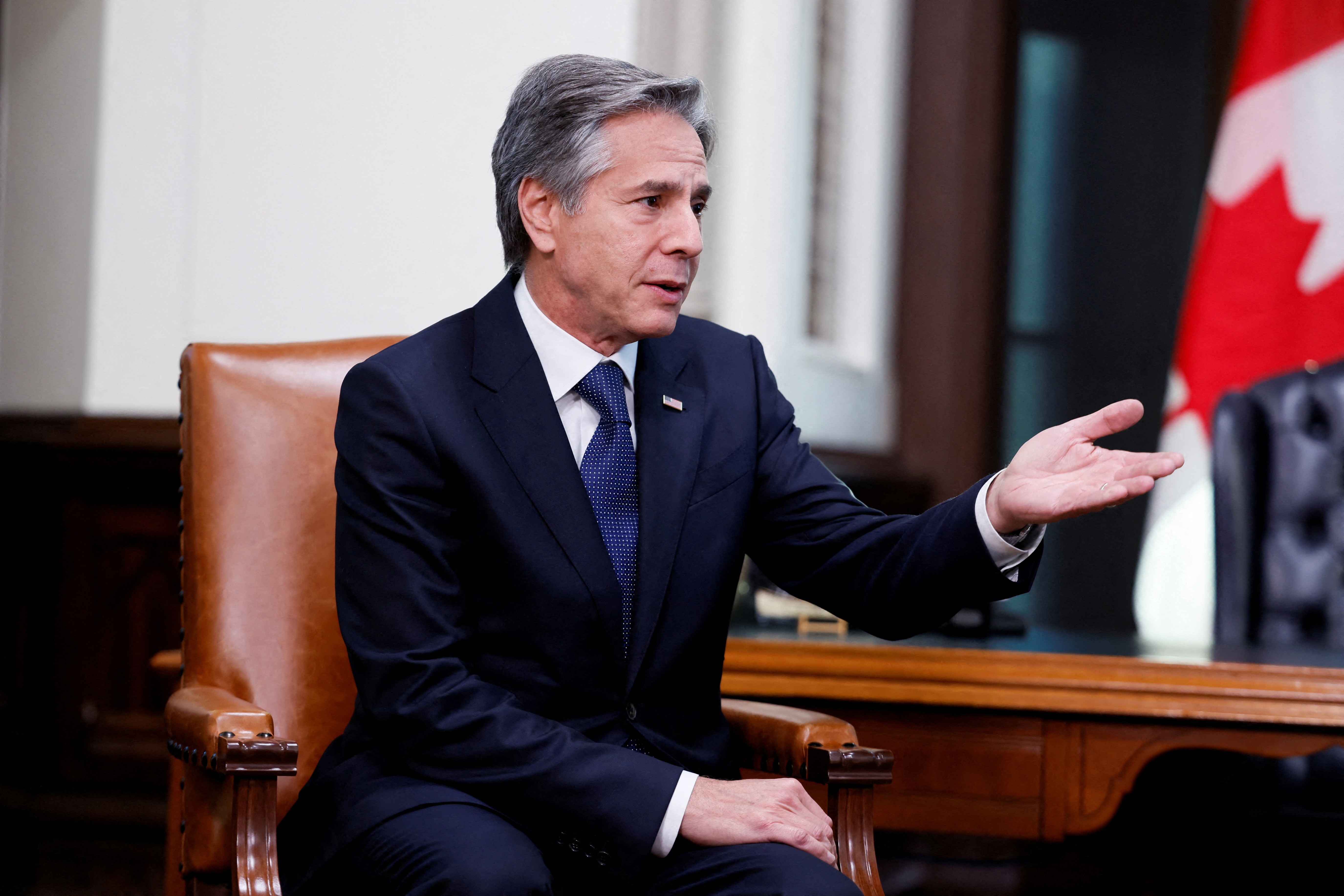 Antony Blinken sits at a wooden desk in a suit and tie, gesturing. A Canadian flag can be seen in the background.