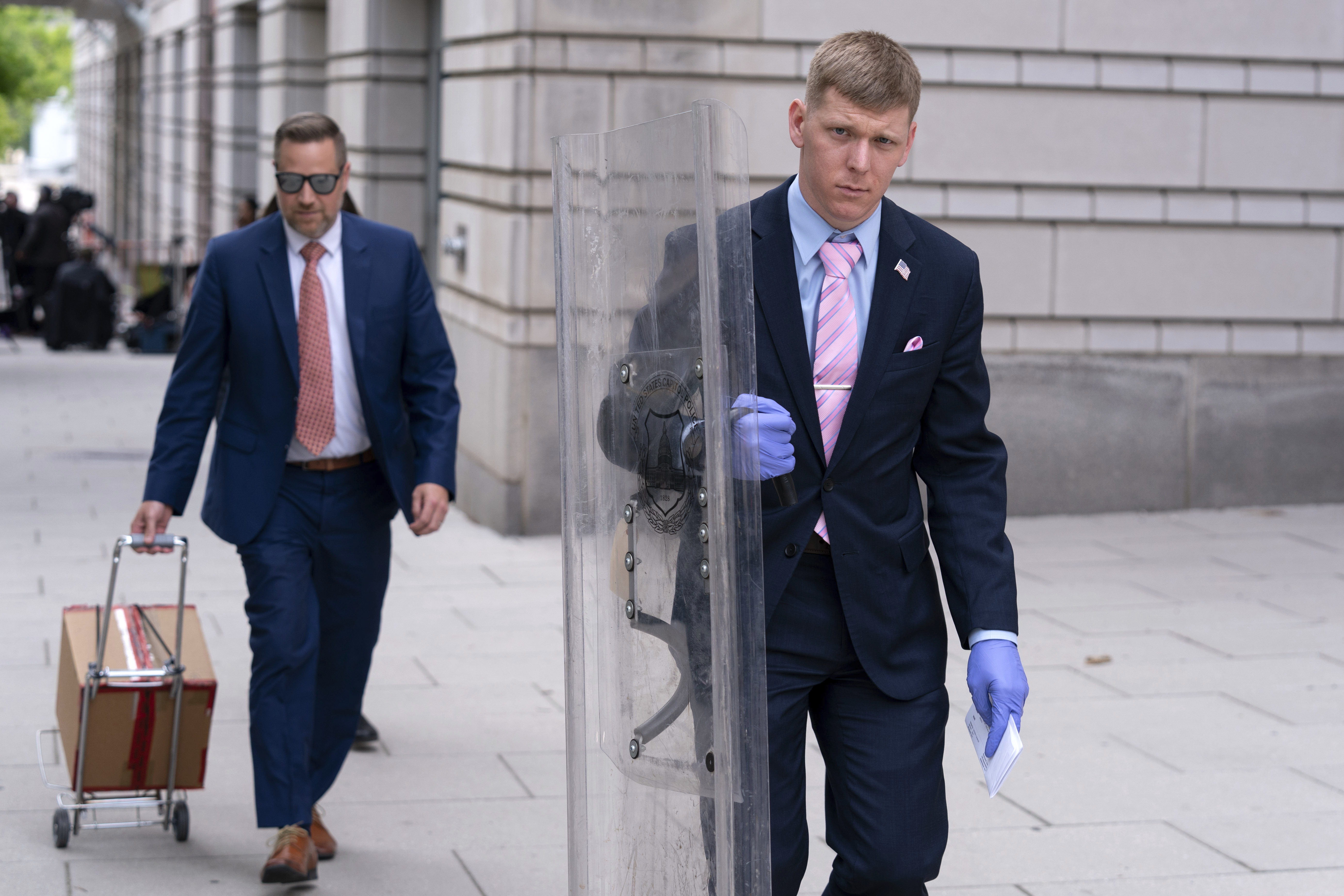 A man wearing blue evidence gloves and a suit and pink tie walks outside a courthouse with a clear plastic riot shield as evidence.