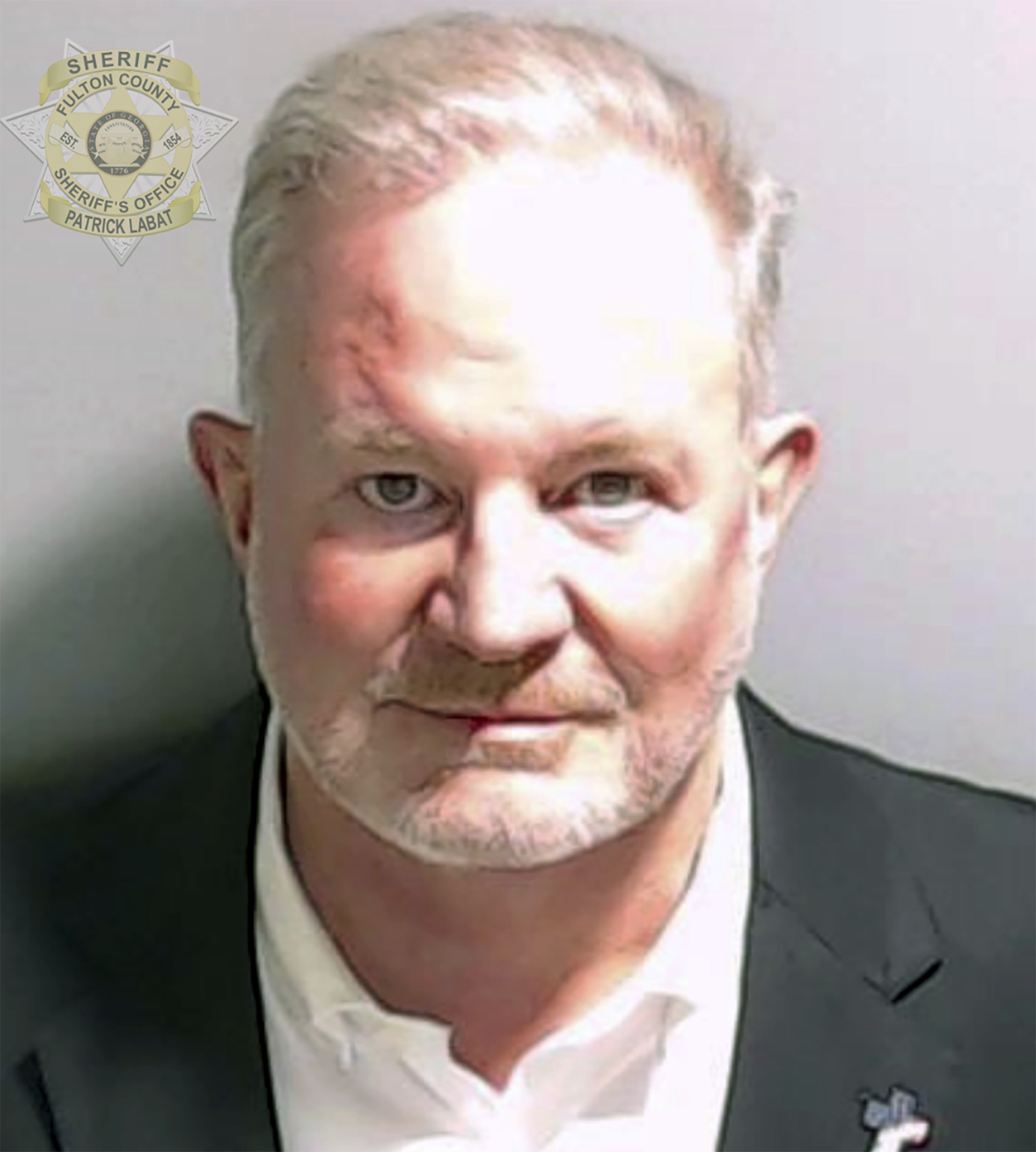 A mugshot of Scott Hall, dressed in a white shirt and dark suit. A Fulton County seal is superimposed at the top of the image.