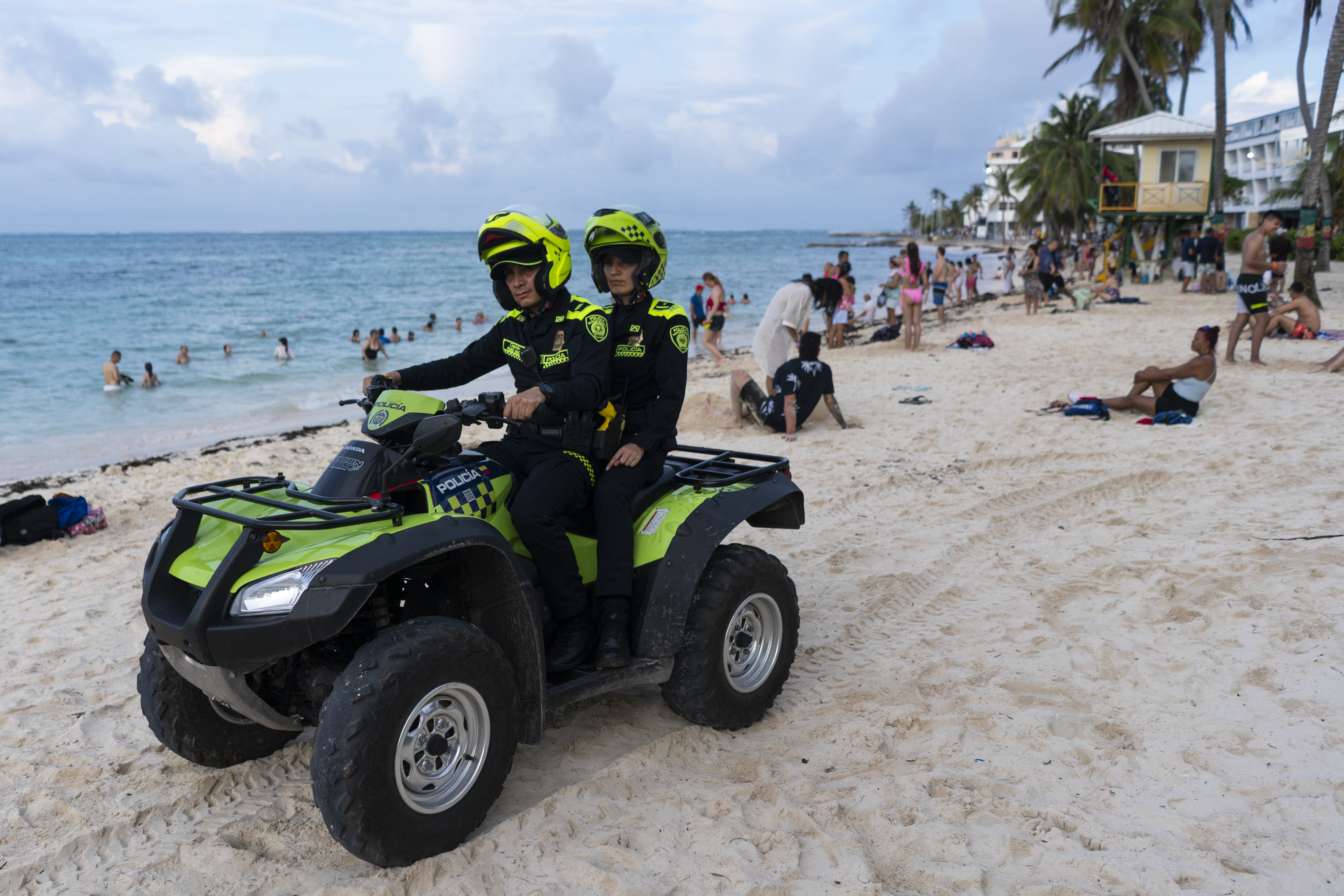 Two police officers ride together on a green ATV, passing across a touristy beach.