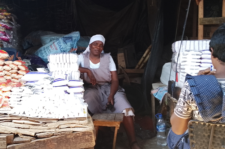 A photo of a person sitting next to a table with piles of grains and spices with another person sitting opposite them.