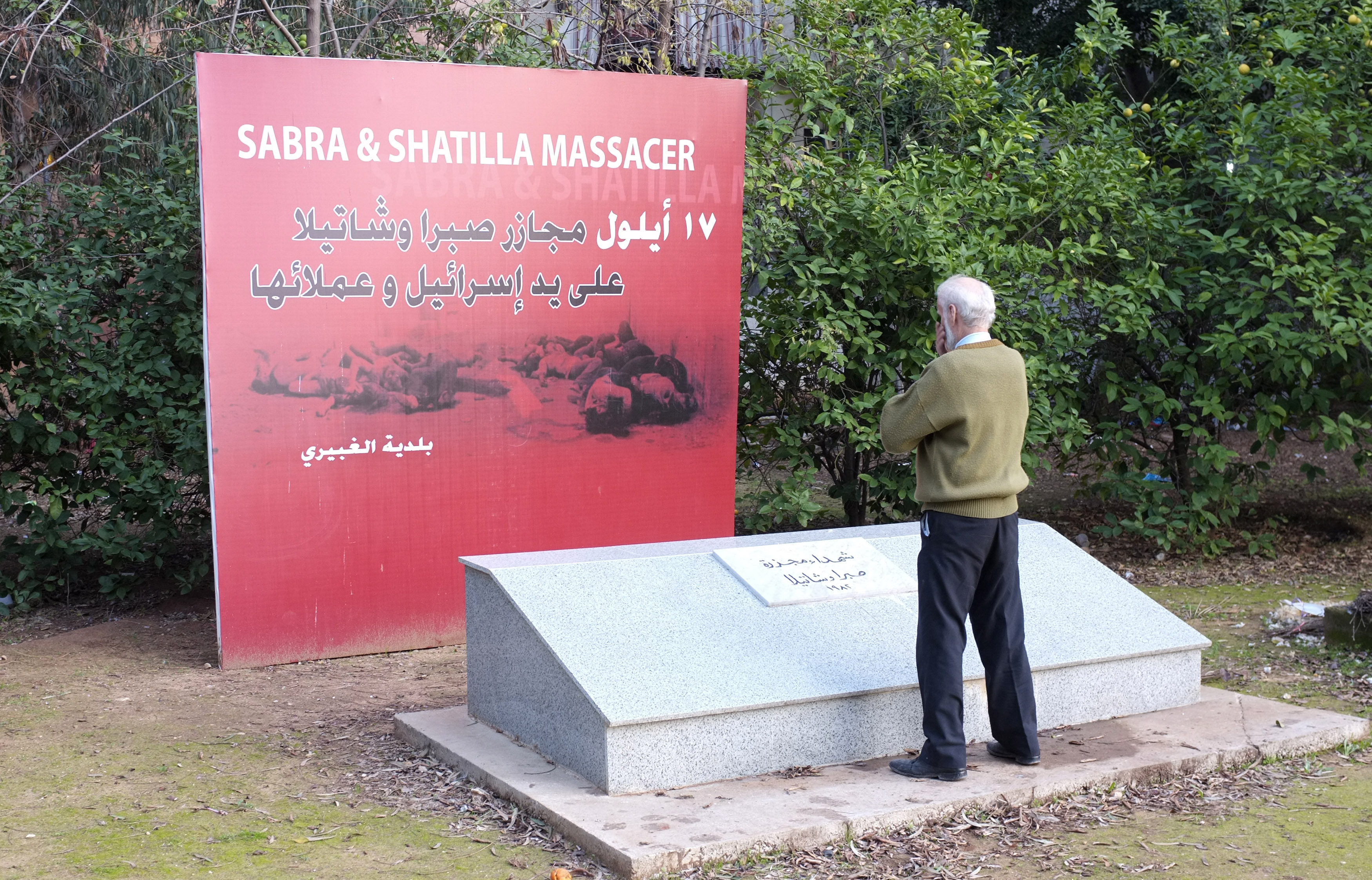 Yousef Hamzeh, a white-haired man seen from behind, stands at a granite memorial. Behind it is a red sign that reads "Sabra and Shatila massacre".