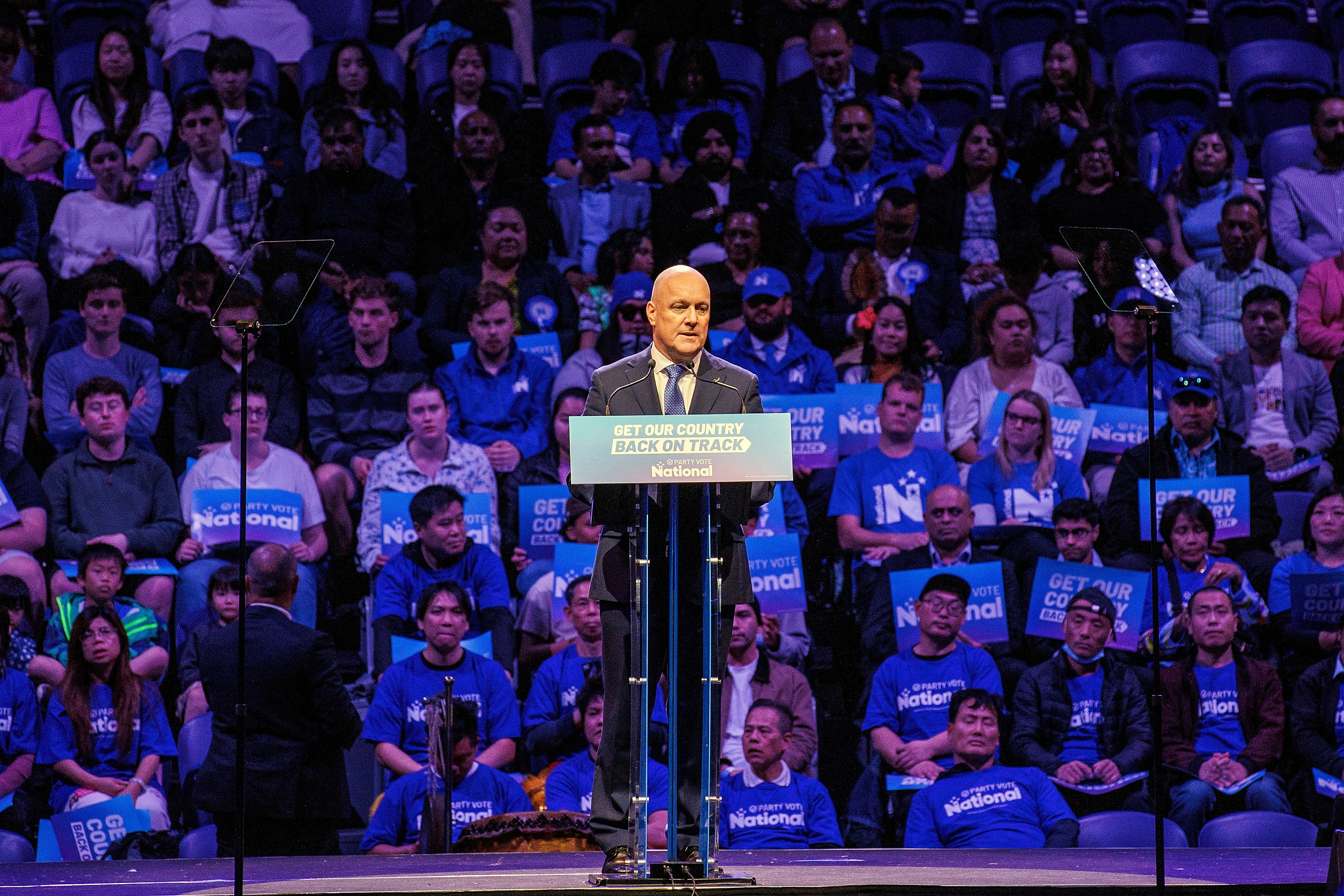 A man in a black suit stands in front of a podium in front of rows of seated supporters illuminated in blue light