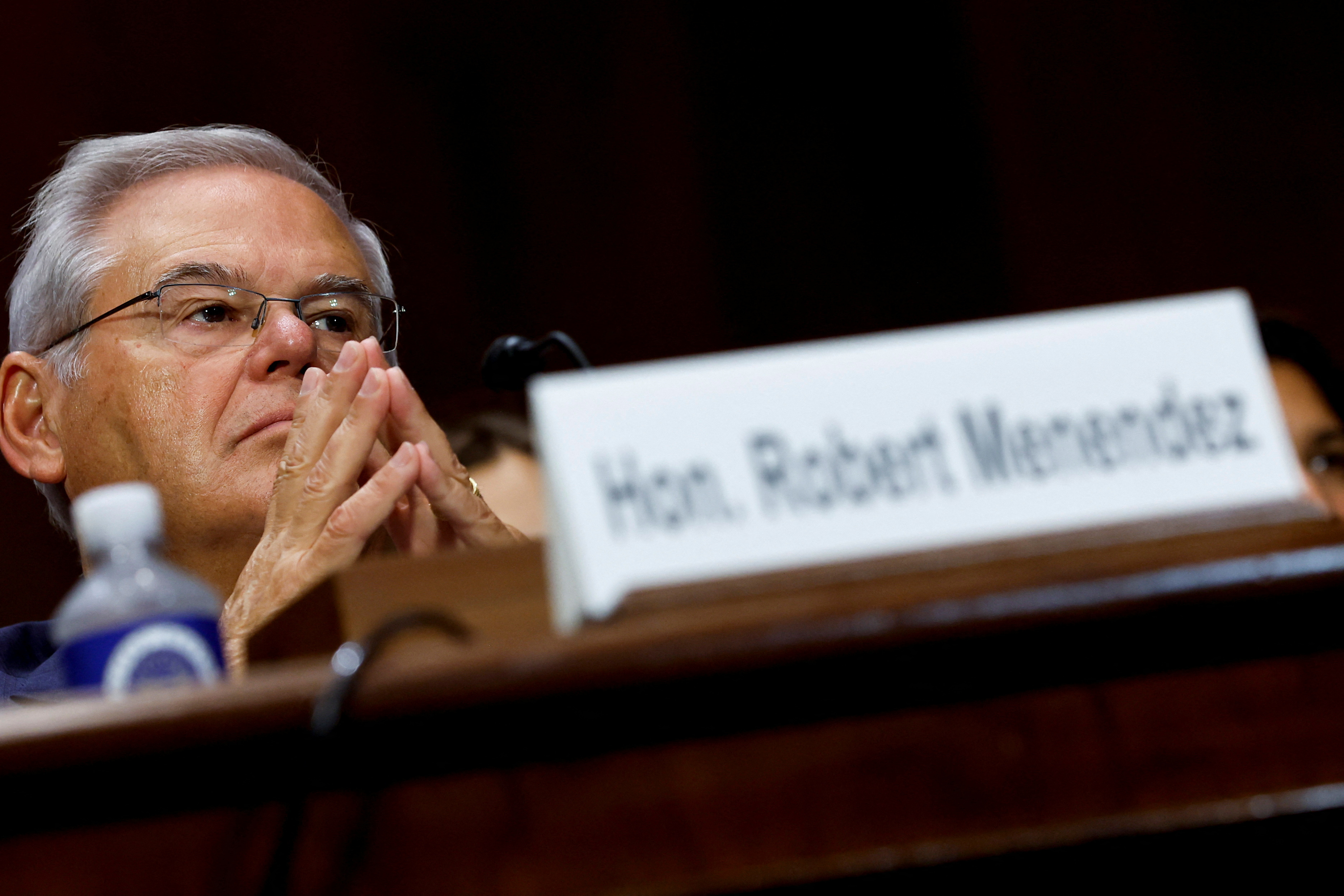 Senator Robert Menendez sits in a committee hearing, his two hands perched together in front of his face and a name tag on the table in front of him.