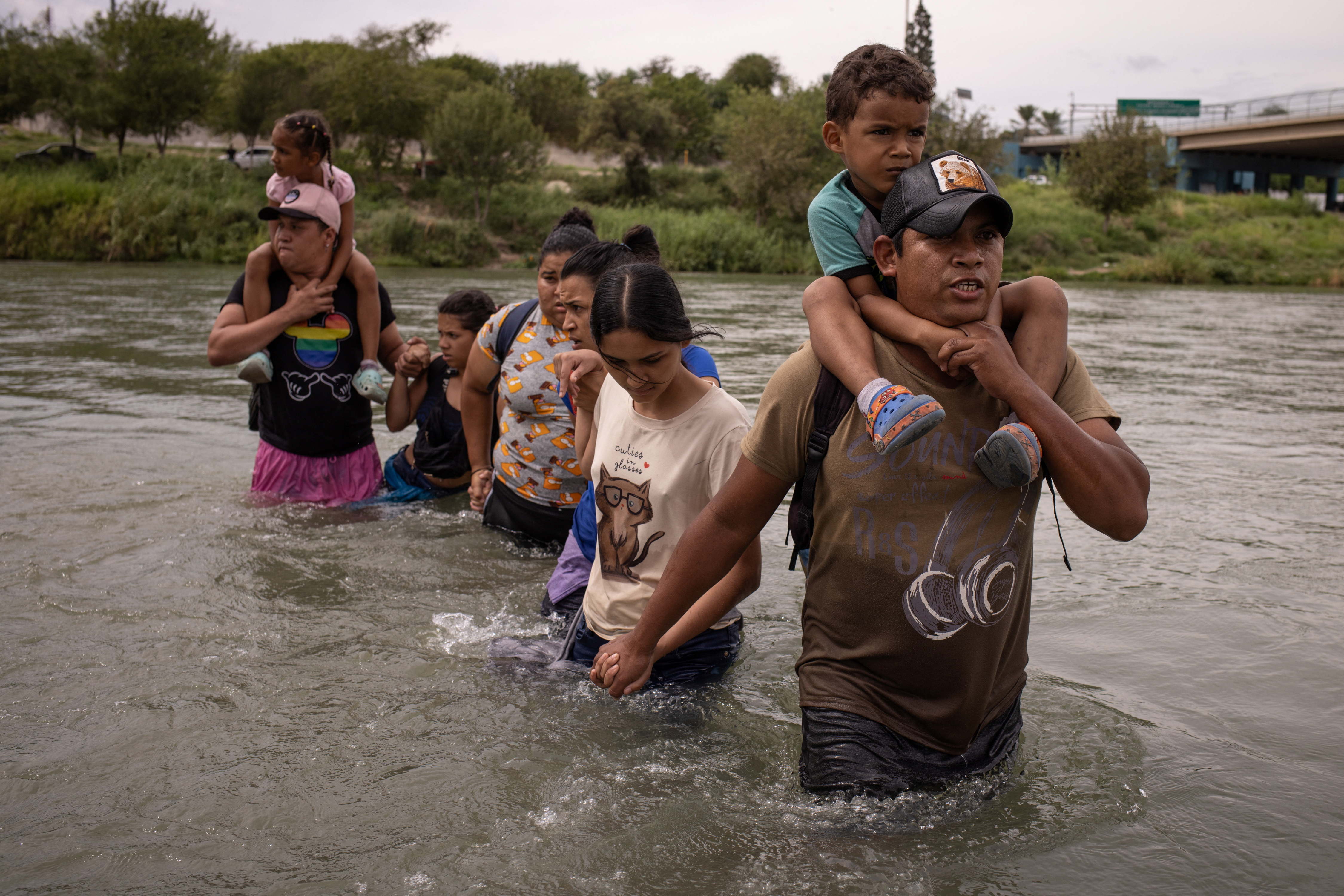 A man carries a four-year-old on his shoulders as he wades through waist-deep river water.