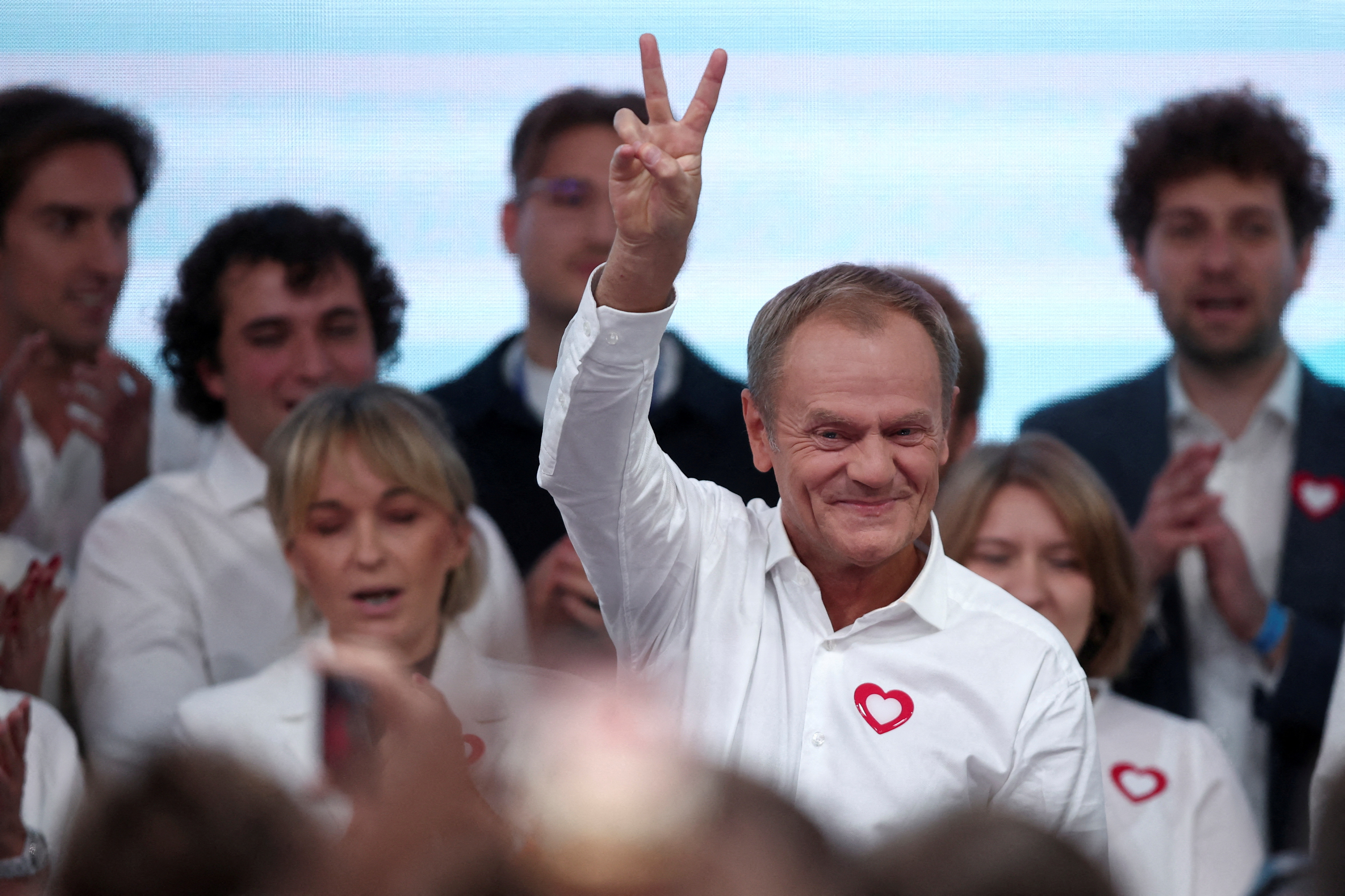 a man in a white shirt with a red heart sticker on his chest holds a peace sign with his fingers with supporters behind him