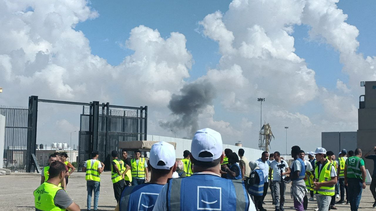 People in blue or neon vests and white caps watch as smoke rises on the horizon at a border crossing.
