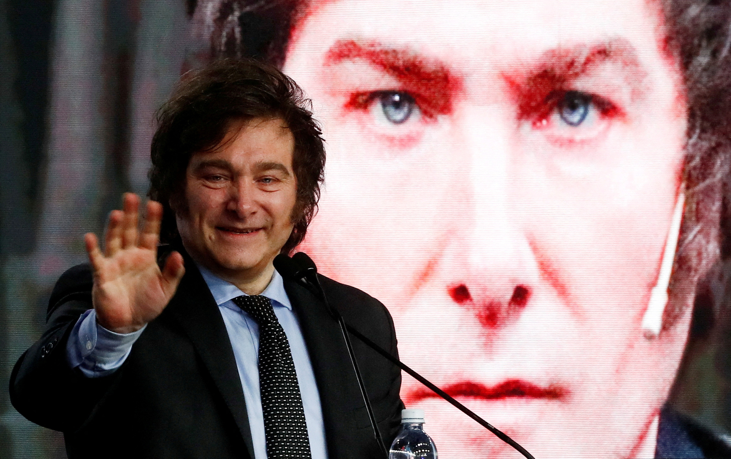 Javier Milei speaks into a microphone and waves. Behind him, a screen displays a giant picture of his face.