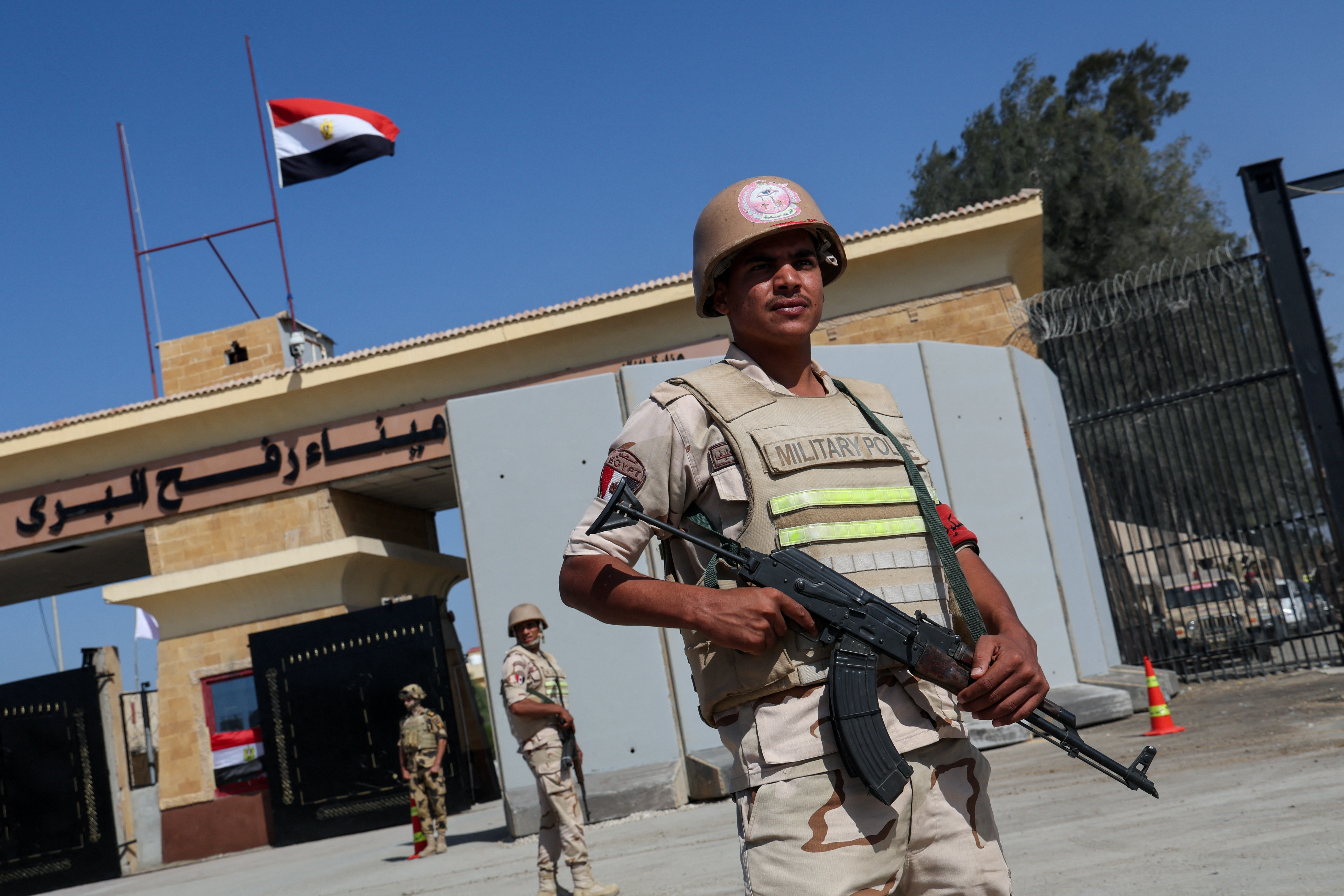 A military man carries a rifle and wears a tan flak jacket, a helmet and short-sleeved fatigues. Behind him, a border crossing building is topped with the Egyptian flag.