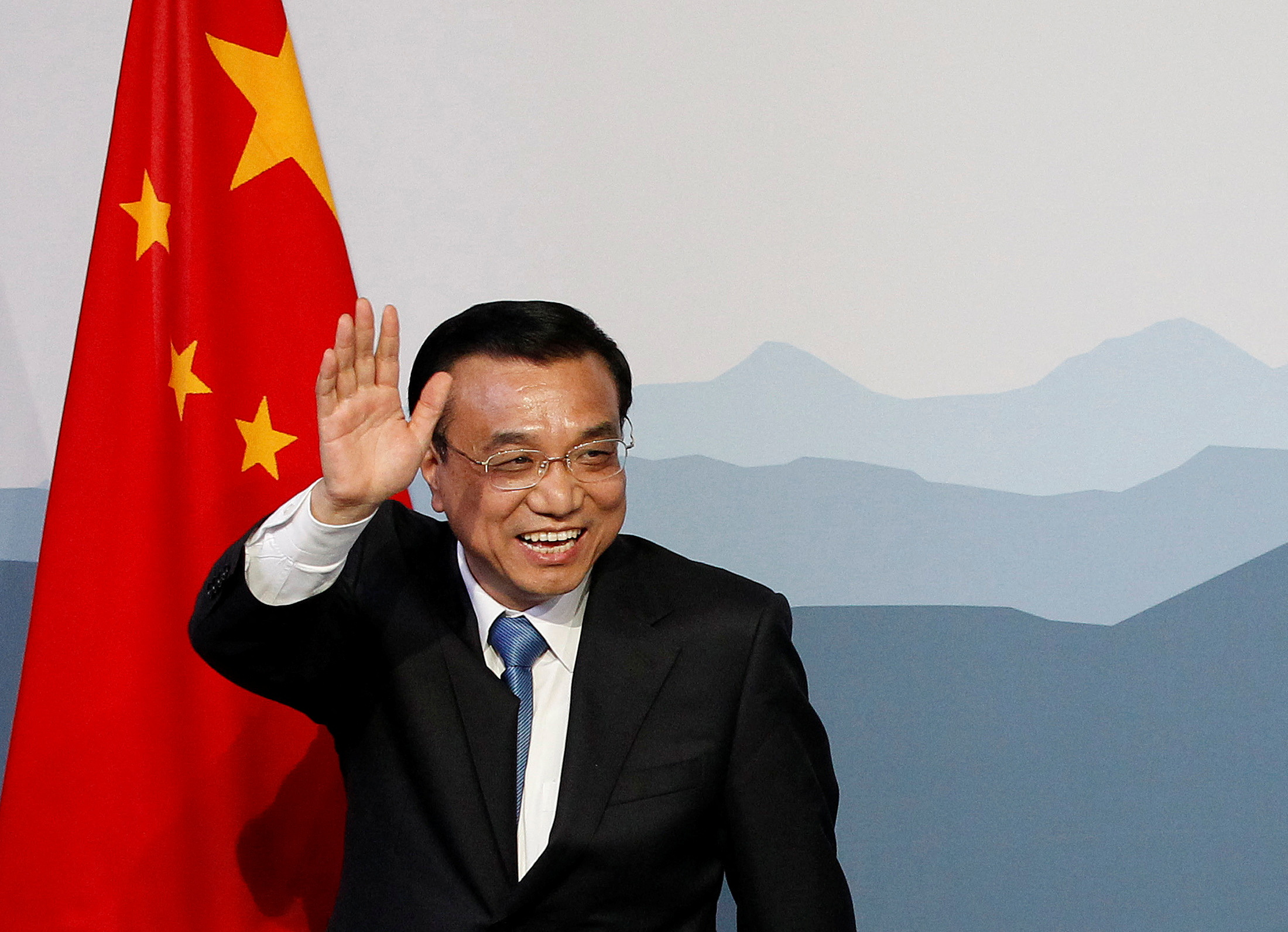 Li Keqiang waving as he walks past a Chinese flag. He is smiling broadly.