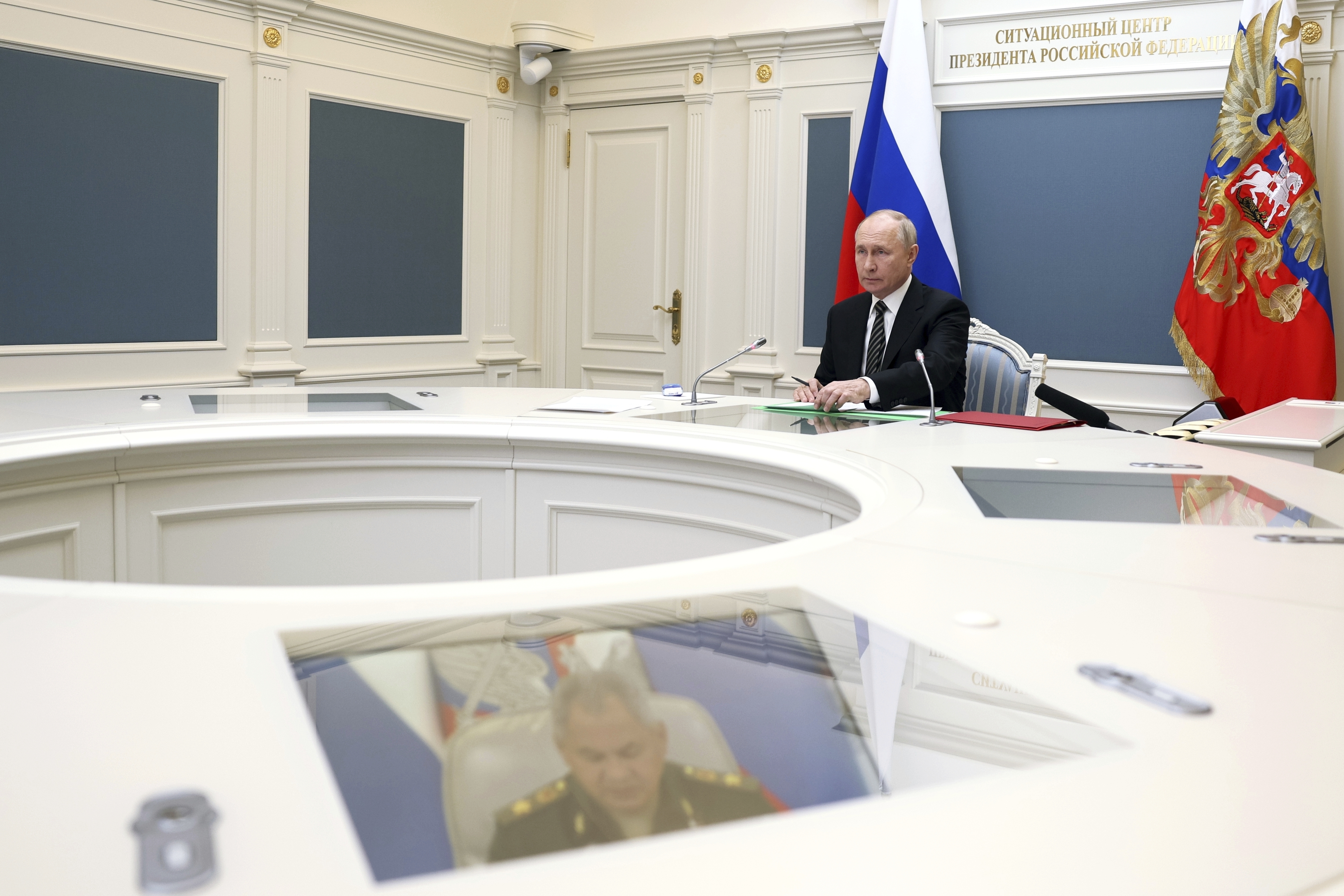 Putin seated at a round table in the Kremlin. The room is white with Russian flags. Defence minister Sergey Shoigu can be seen on a TV screen embedded in the table.