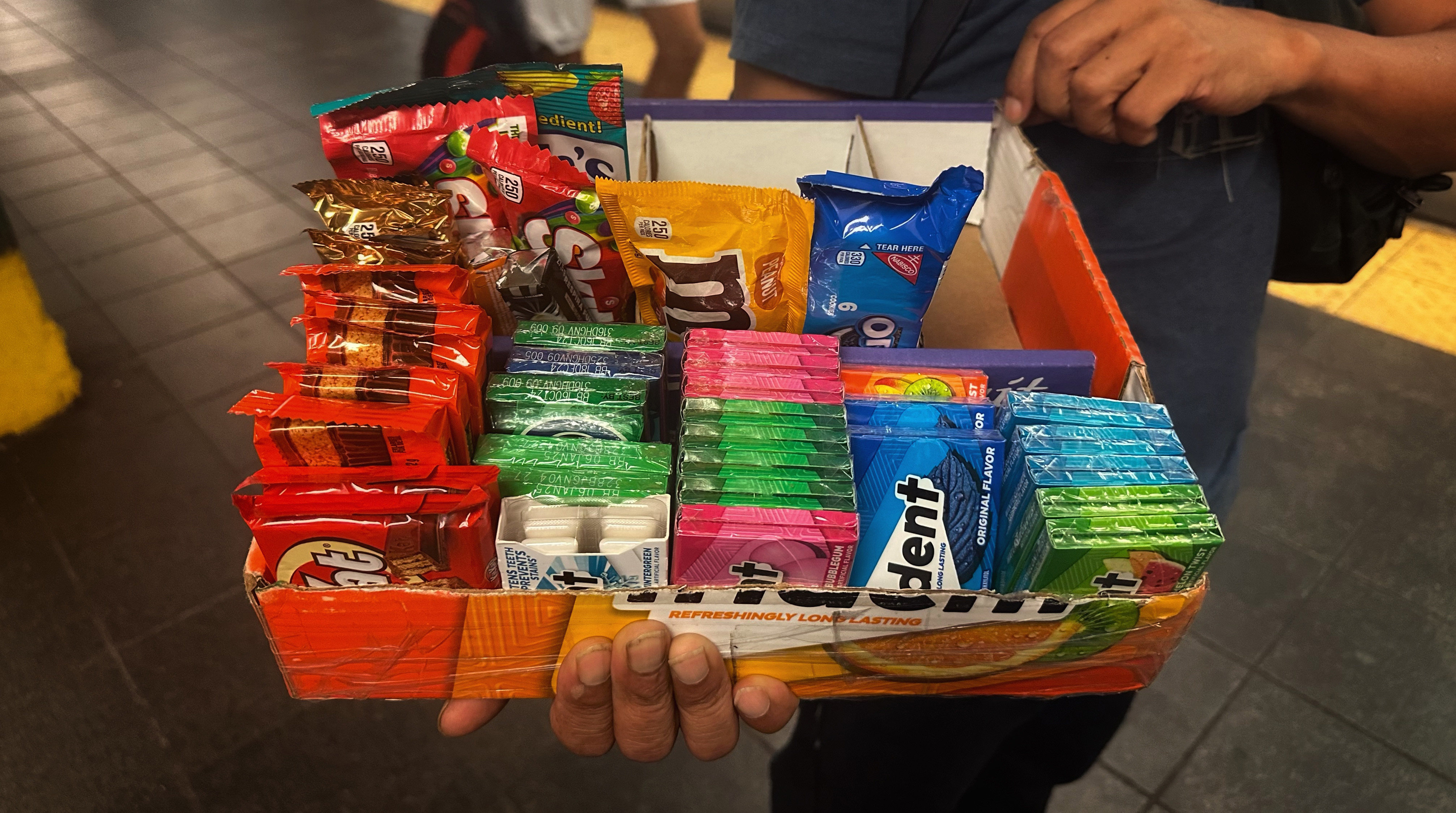 A person holds out a tray of candy, with Trident gum, Kit Kats and M&Ms.