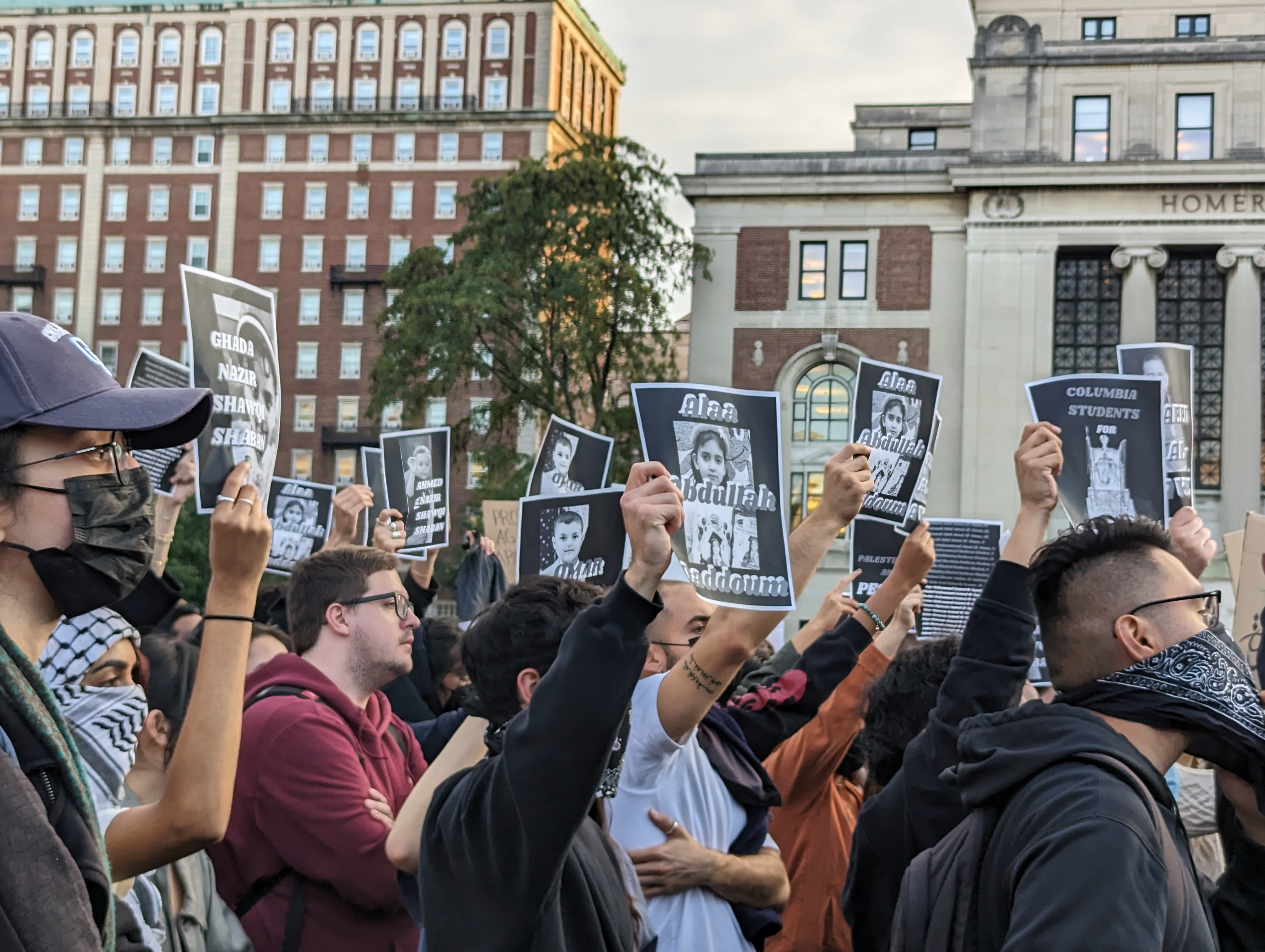 A packed crowd of students raise black-and-white print-outs into the air, featuring the faces of small children. Some of the students where hygienic masks or bandanas over their faces.