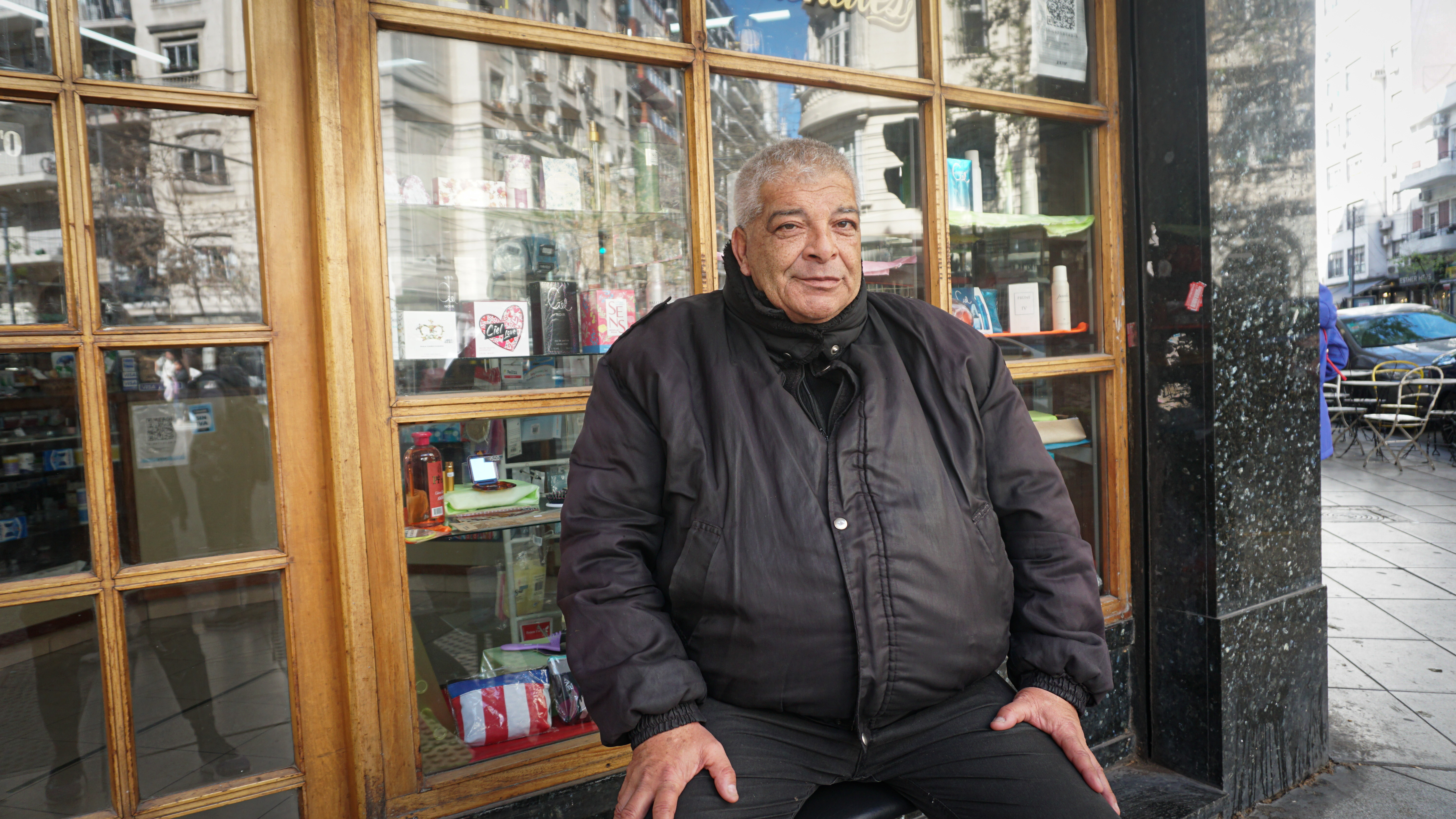 A man in a black jacket sits on a window ledge on the street: Behind him, a store display is visible behind the glass.