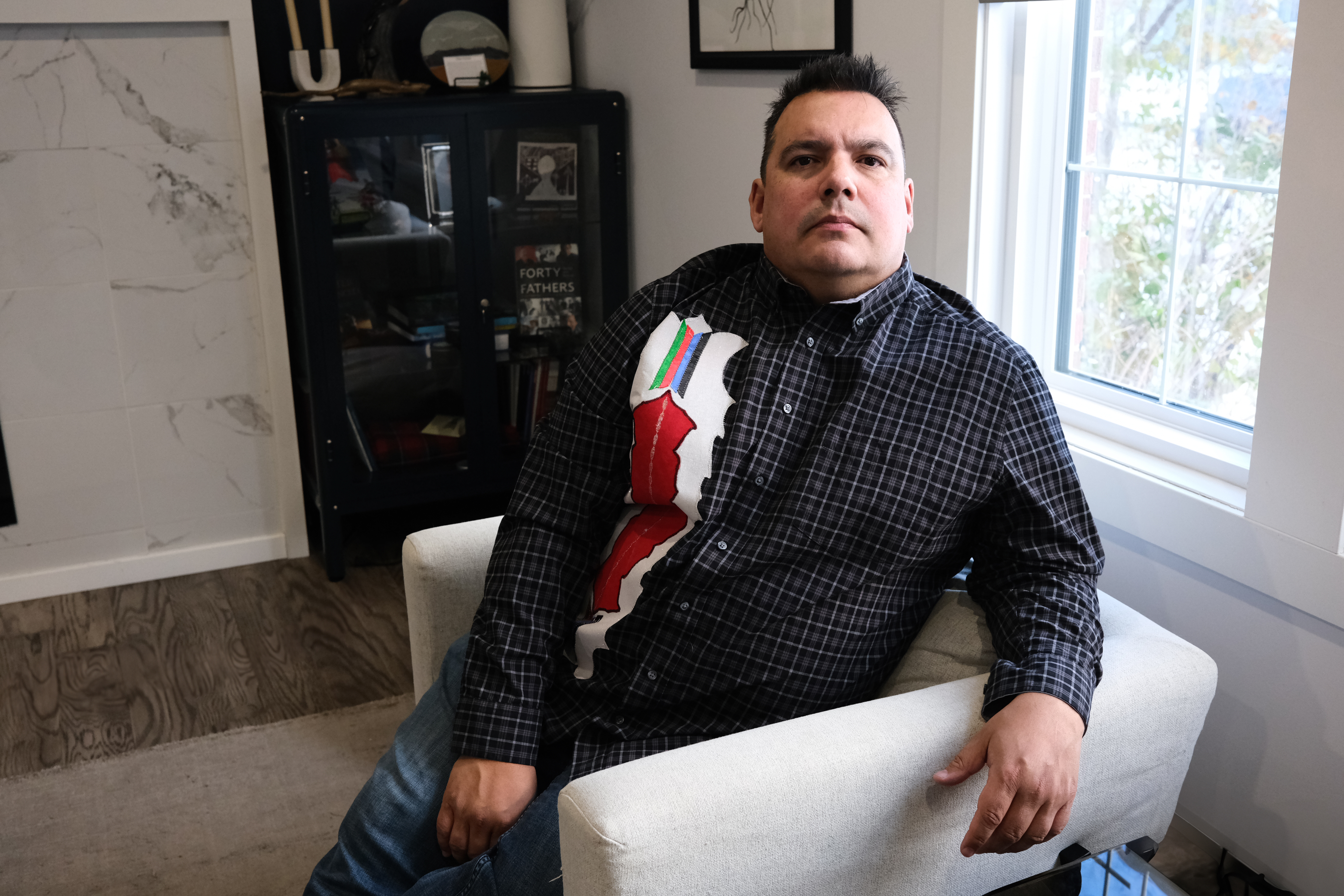 Niigaan Sinclair sits in a beige chair in a living area, with a book shelf visible behind him.
